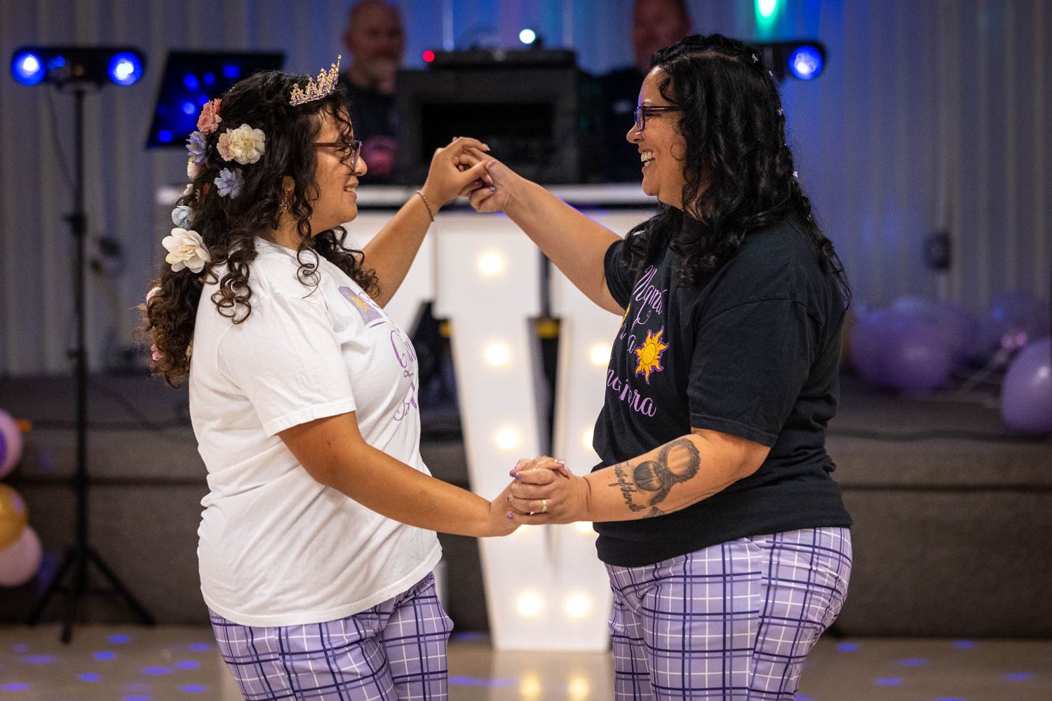 Two women joyfully dance together holding hands at a quinceañera celebration with festive lighting in background.