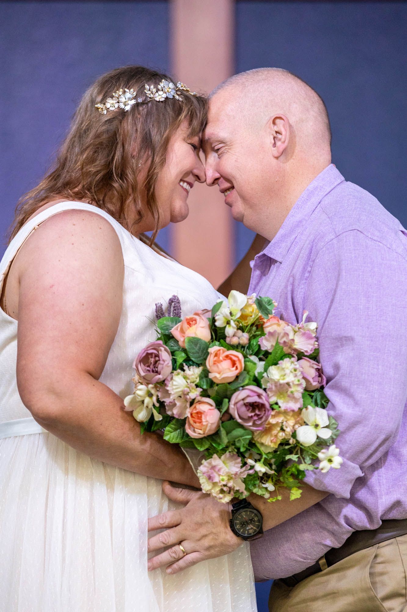Smiling bride and groom touch foreheads tenderly, bride holding pastel rose bouquet in church ceremony.