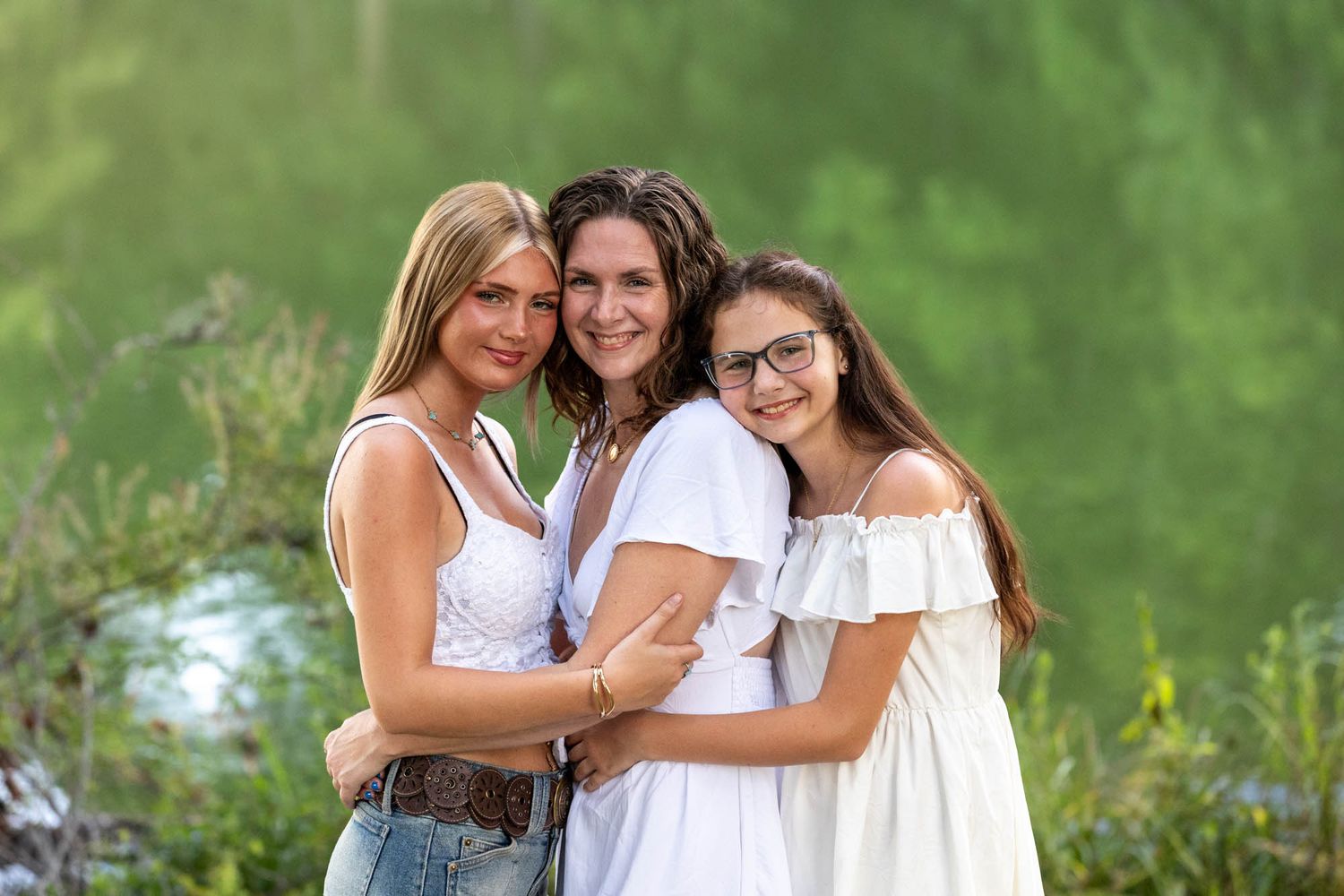 Three smiling women pose together outdoors, wearing white tops, with lush green trees in the background.