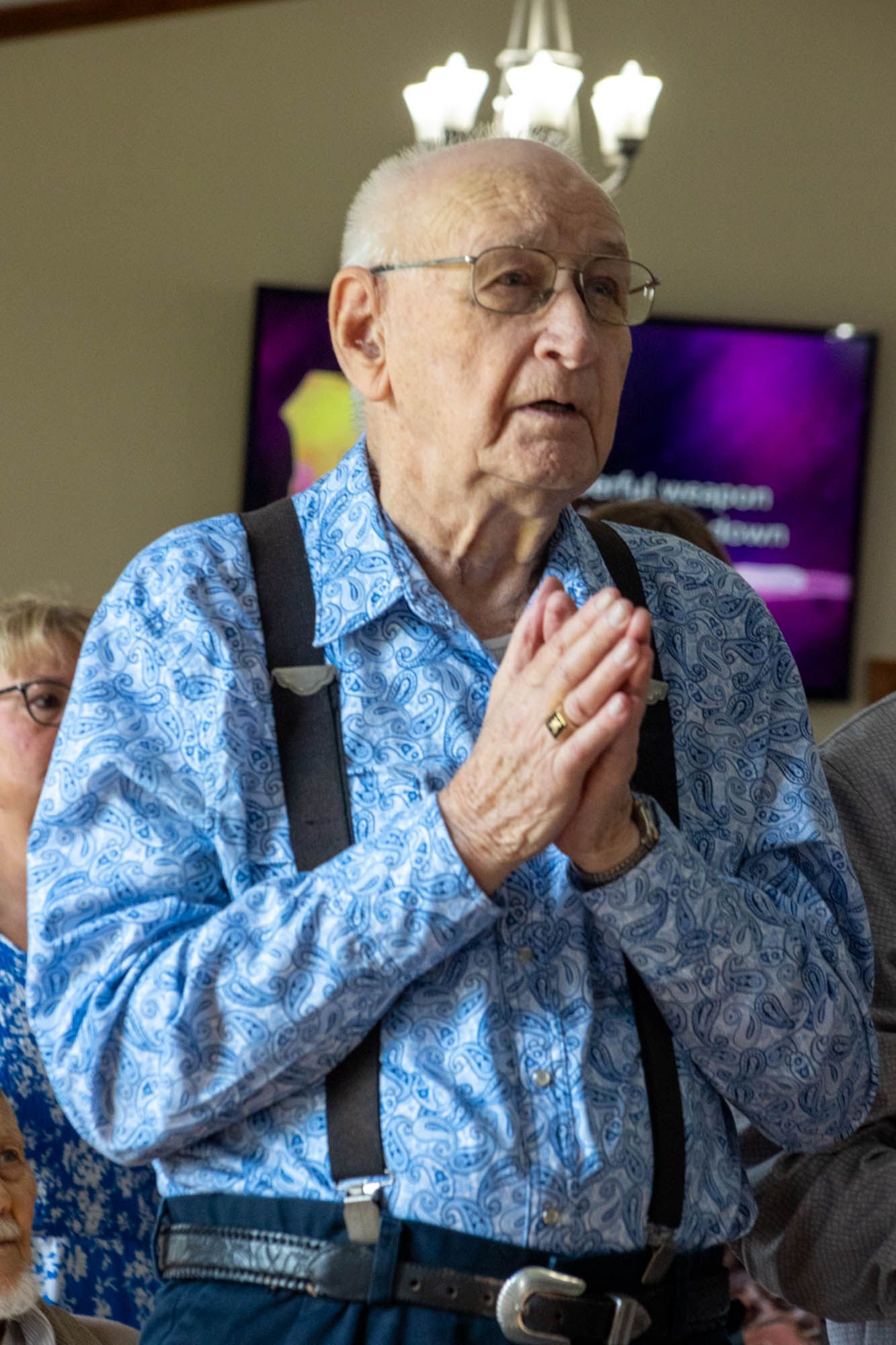 Elderly man in blue floral shirt with suspenders praying with hands clasped at church service.