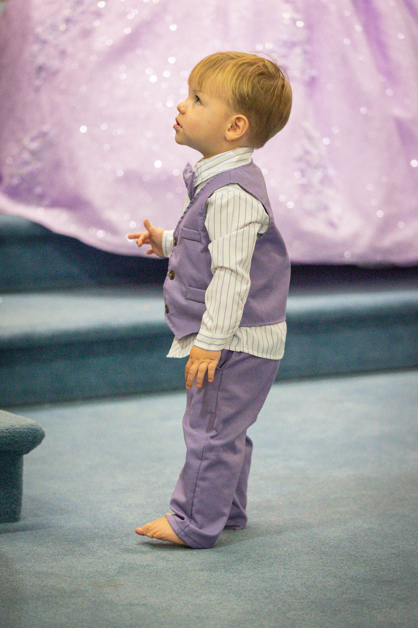 Toddler boy in purple vest suit standing barefoot at quinceañera party.