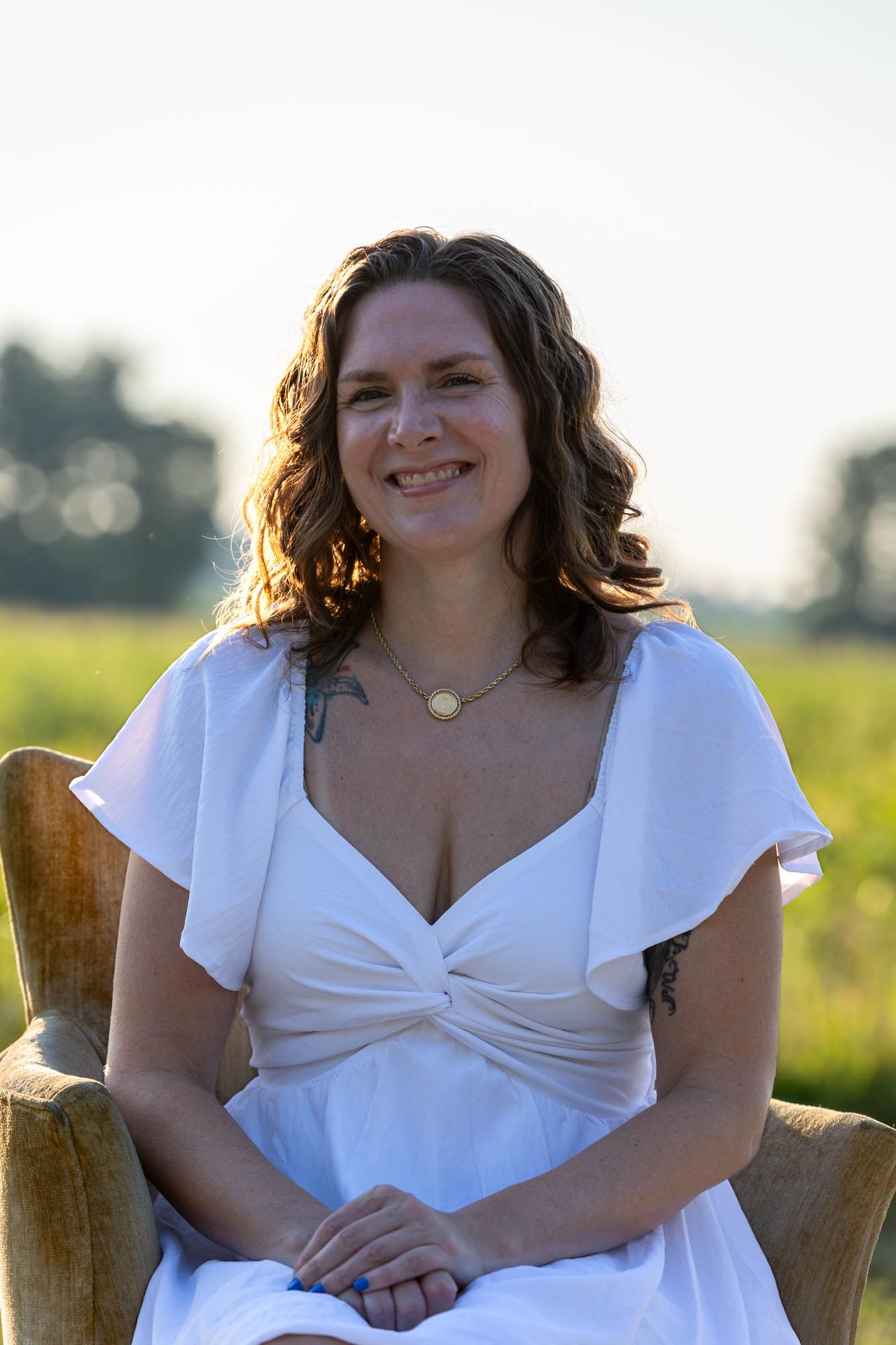 Smiling woman in white dress seated outdoors in golden hour light with green fields in background.