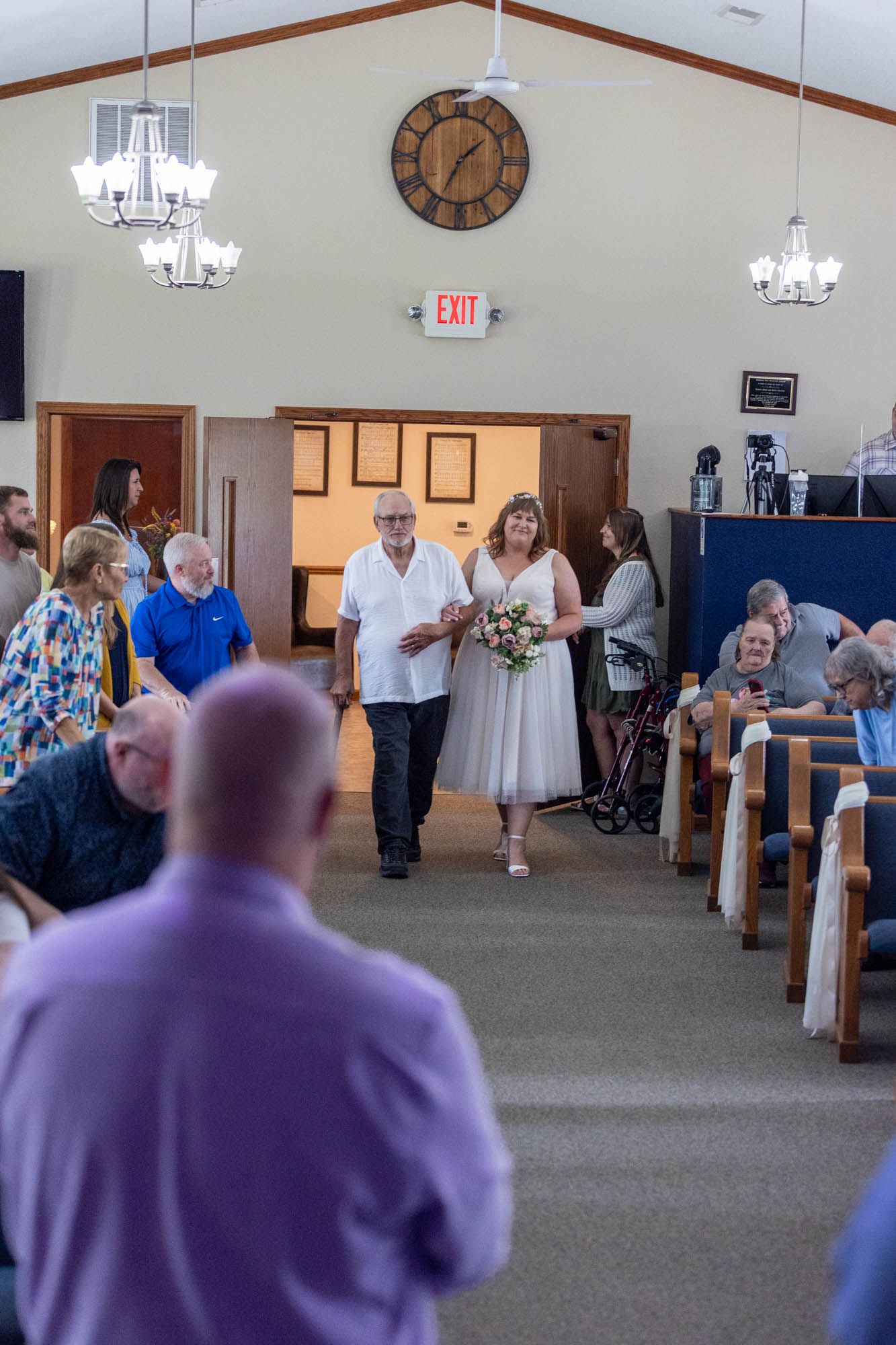 Bride walks down church aisle with escort during wedding ceremony, guests seated in pews, chandelier lighting overhead.