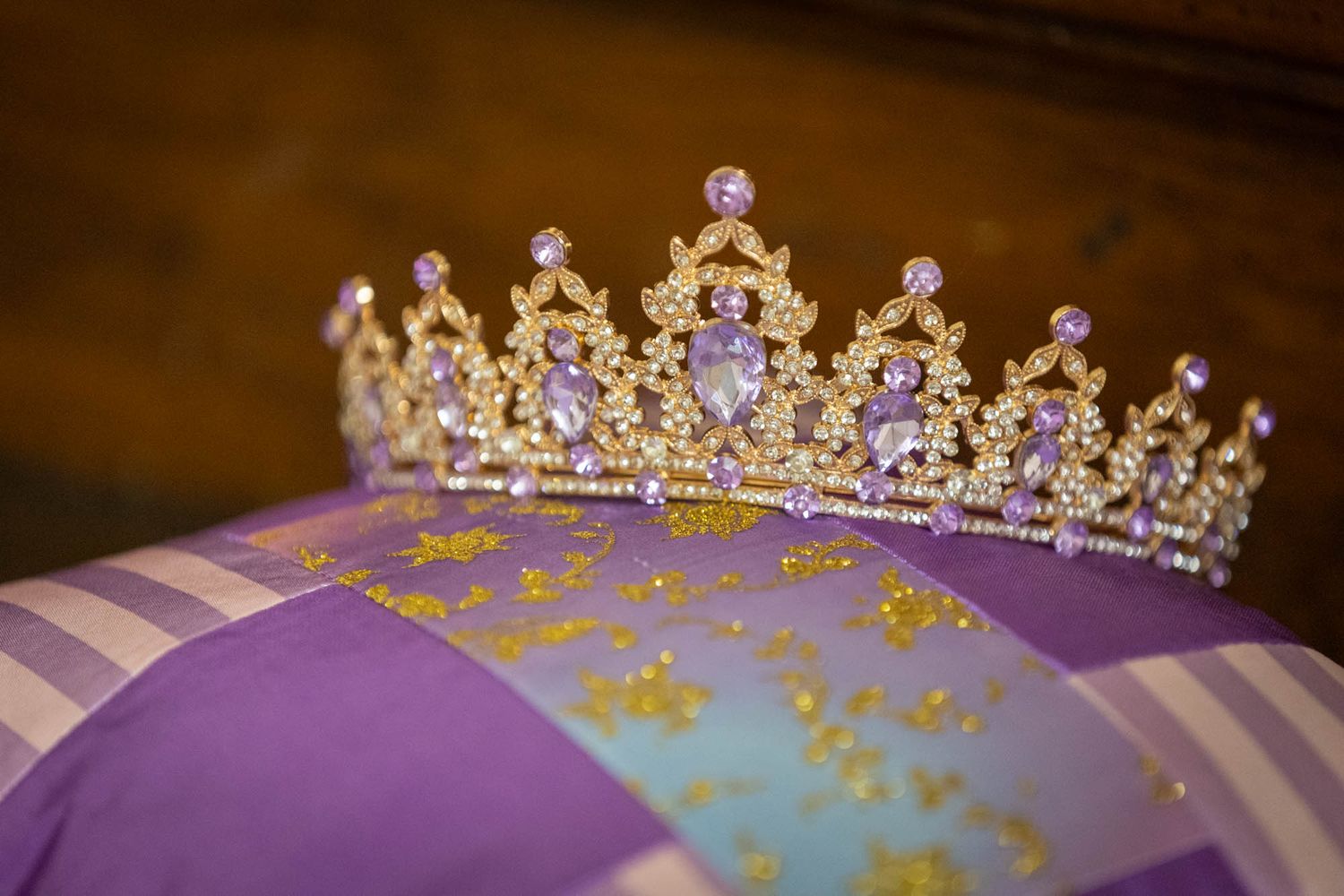 Gold tiara with purple gemstones resting on a lavender silk pillow with gold embroidery.