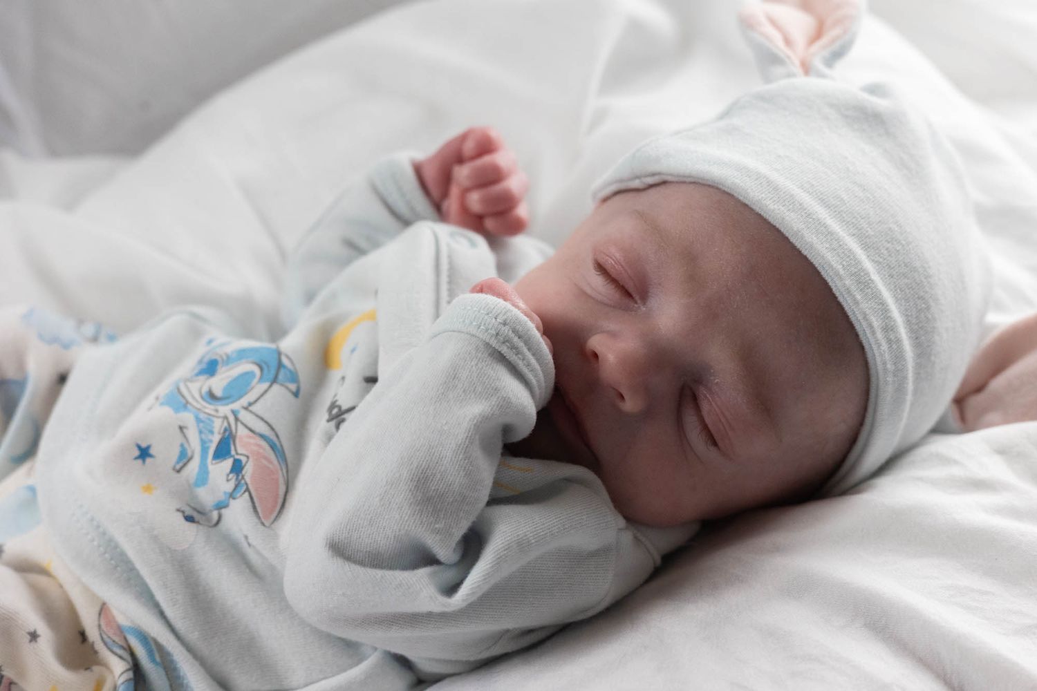 Peaceful newborn baby sleeping in white hat and blue outfit on white bedding.