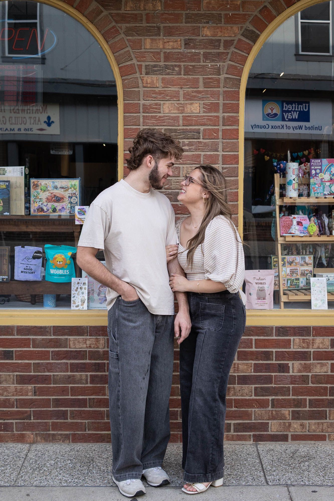 A couple gazes lovingly at each other while standing together in front of a brick building storefront.