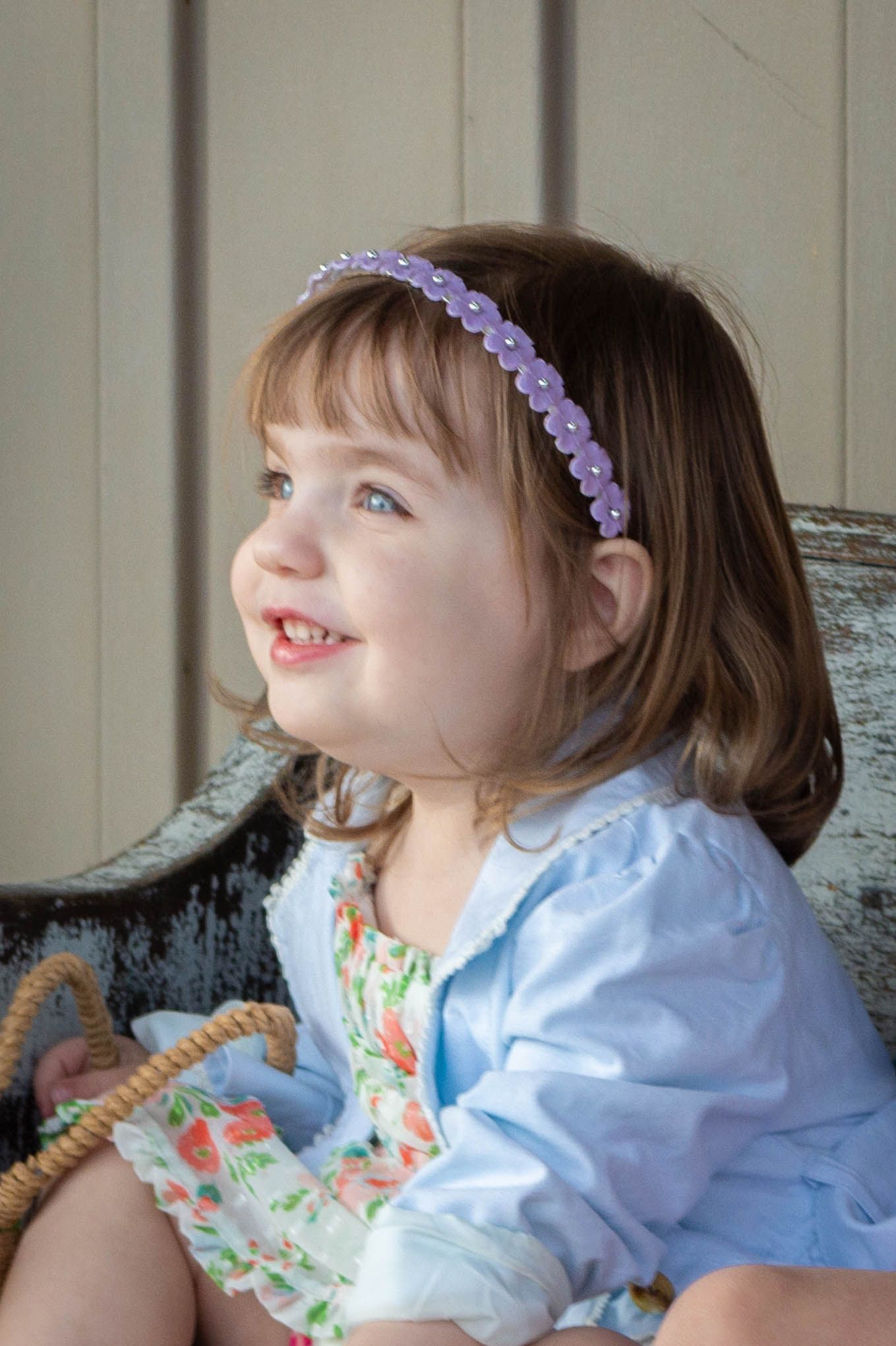 Adorable toddler girl with purple beaded headband smiling while sitting on a vintage bench outdoors.