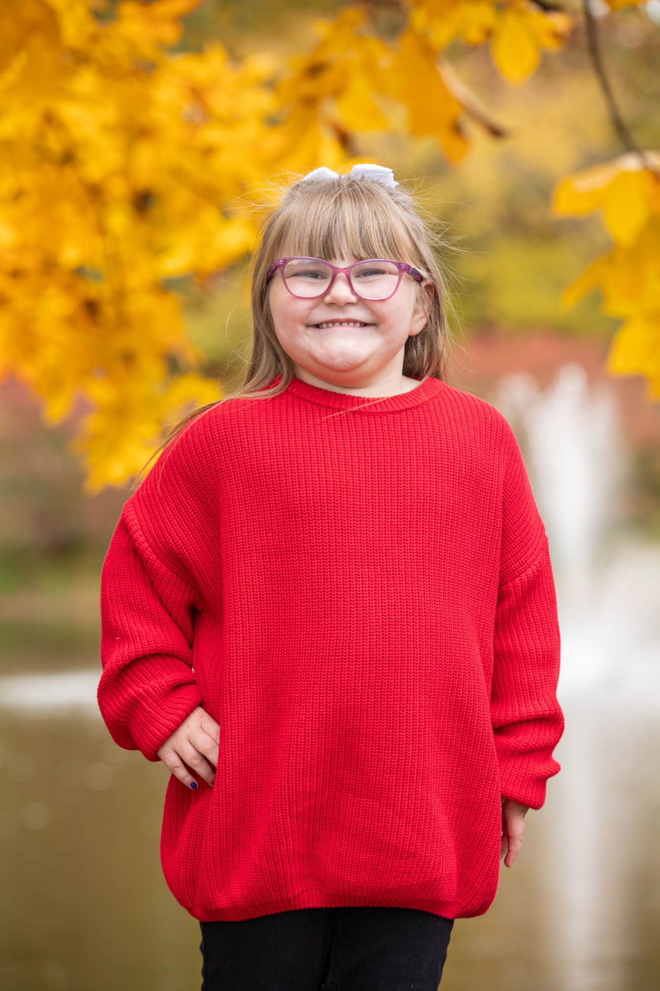 Happy young blonde girl wearing pink glasses and a red sweater smiling in front of golden autumn leaves.