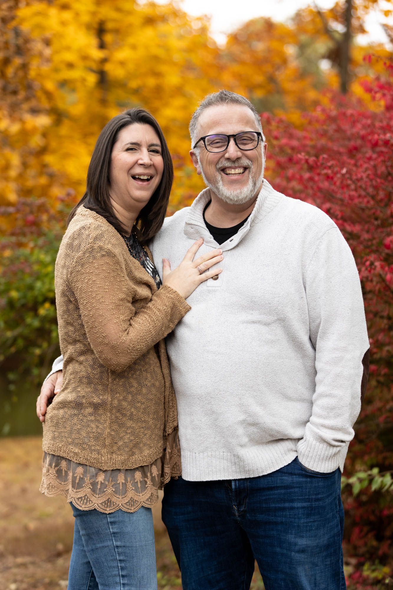 A smiling middle-aged couple laughs together outdoors surrounded by colorful fall foliage.