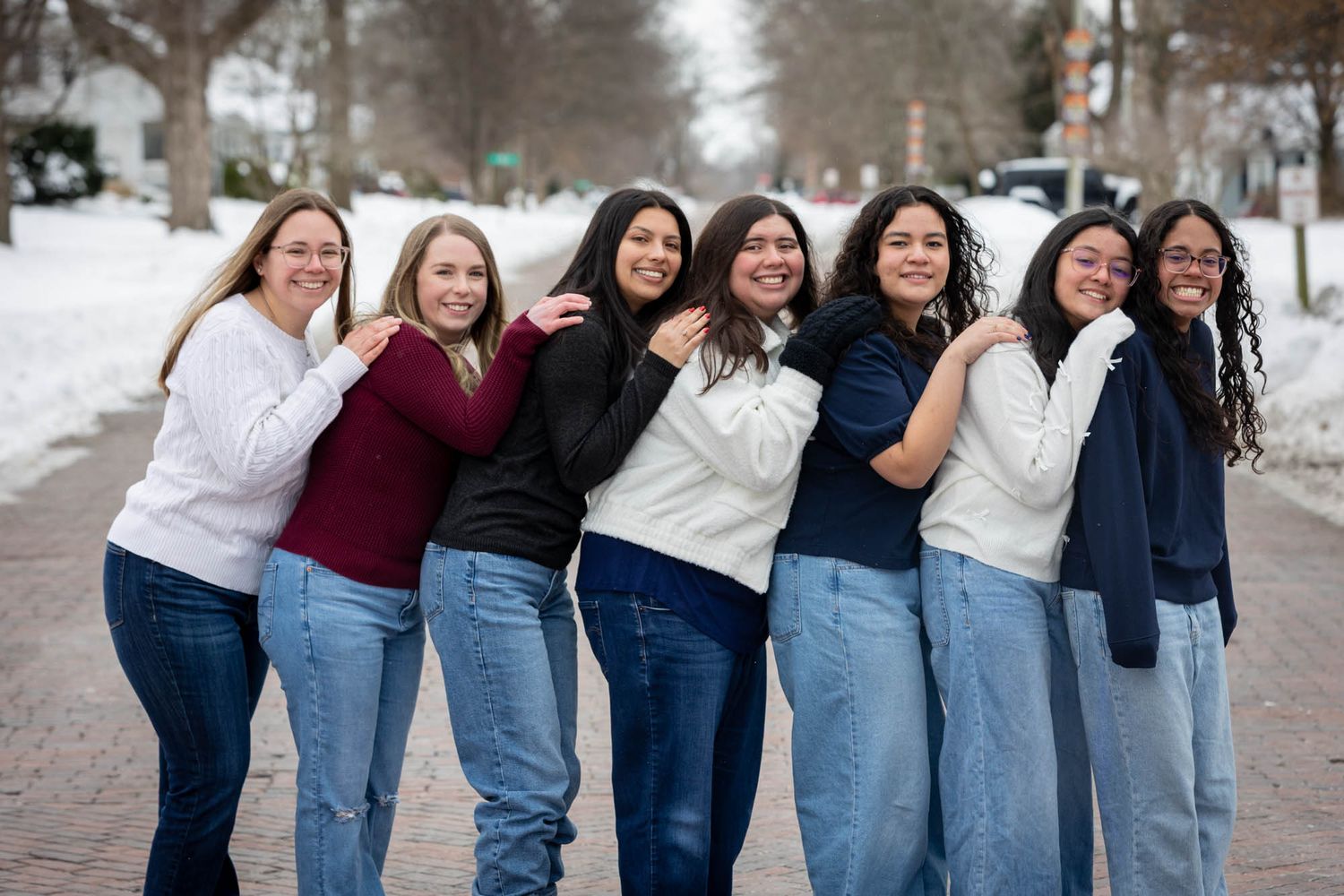 Seven diverse young women in casual sweaters and jeans pose playfully together in winter snow.