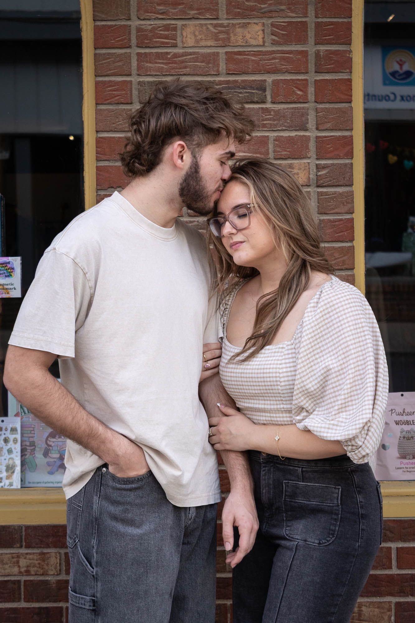 A young couple shares a tender moment, with a man kissing a woman's forehead near a brick wall storefront.