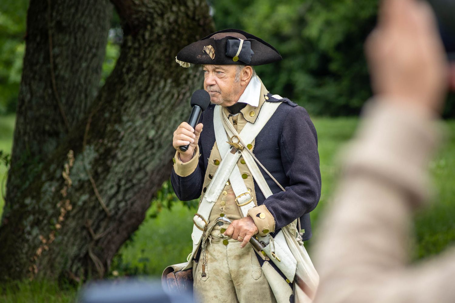 A man in colonial American Revolutionary War uniform speaks outdoors at a historical reenactment event.
