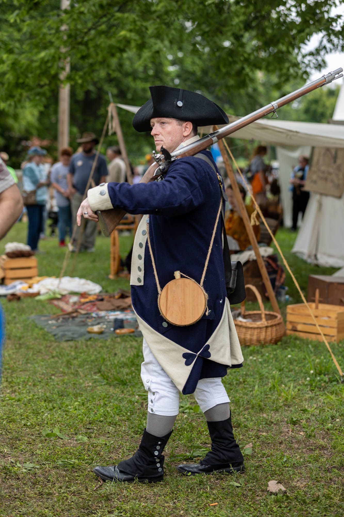 A man in 18th-century colonial military uniform carries a musket at an outdoor historical reenactment event.