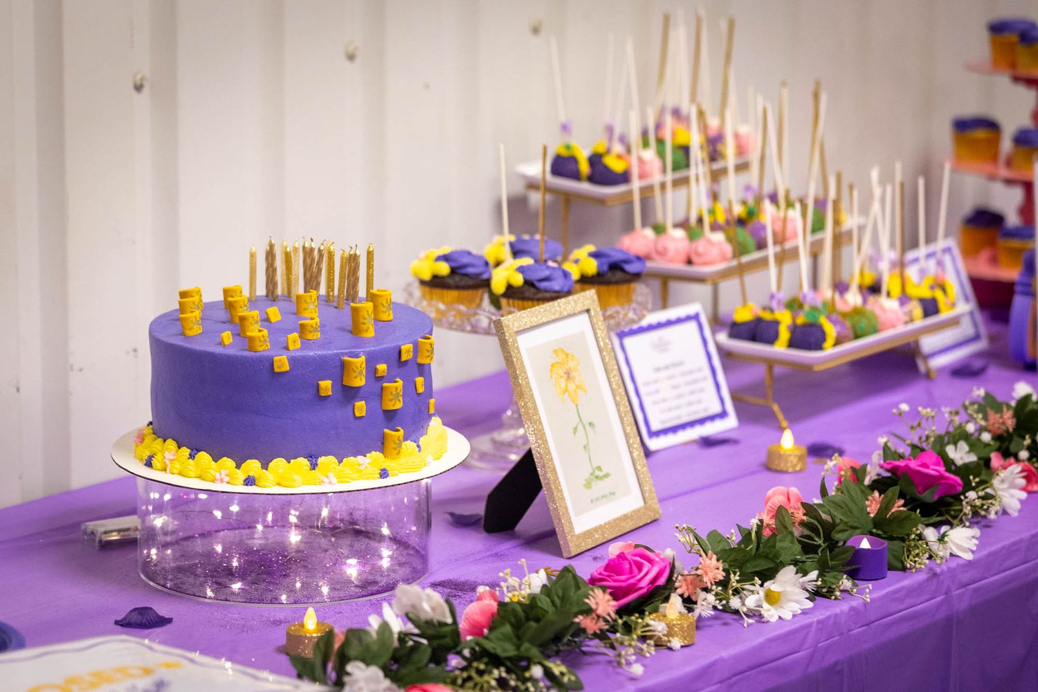 Elegant purple quinceañera dessert table featuring a purple cake with gold candles, cake pops, and floral decorations.
