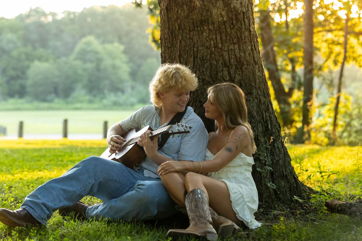 Young couple sitting under a tree, man playing acoustic guitar for woman in white dress and cowboy boots.