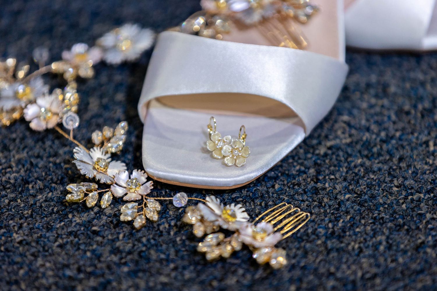 Elegant bridal accessories including gold floral hair comb, crystal jewelry, and white satin wedding heel on dark surface.