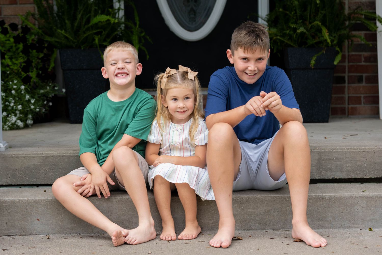 Three smiling children sitting barefoot on front porch steps, two boys in green and blue shirts with a girl in white dress.
