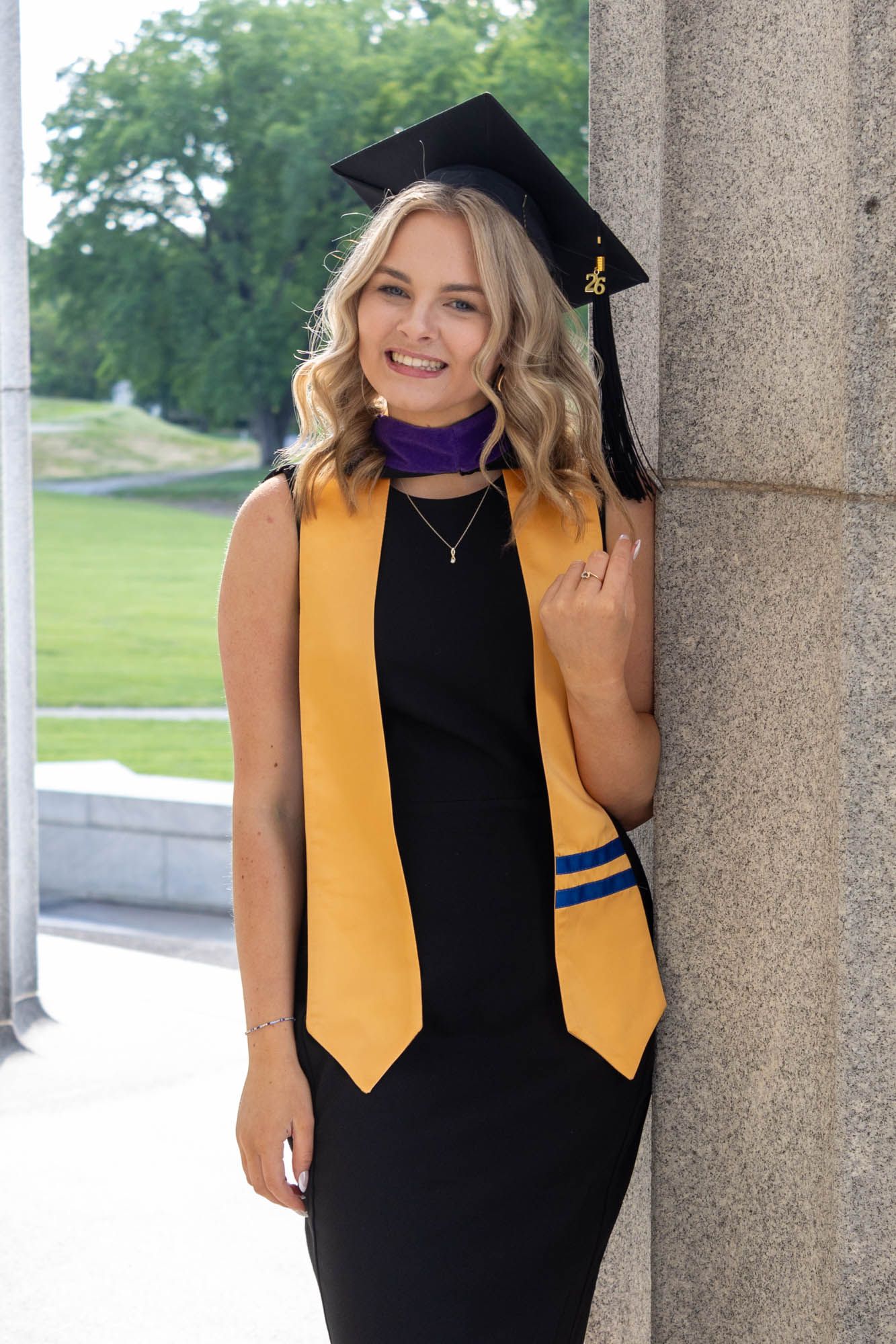 Smiling female graduate in black dress, gold stole, purple hood, and mortarboard cap leaning against stone column outdoors.