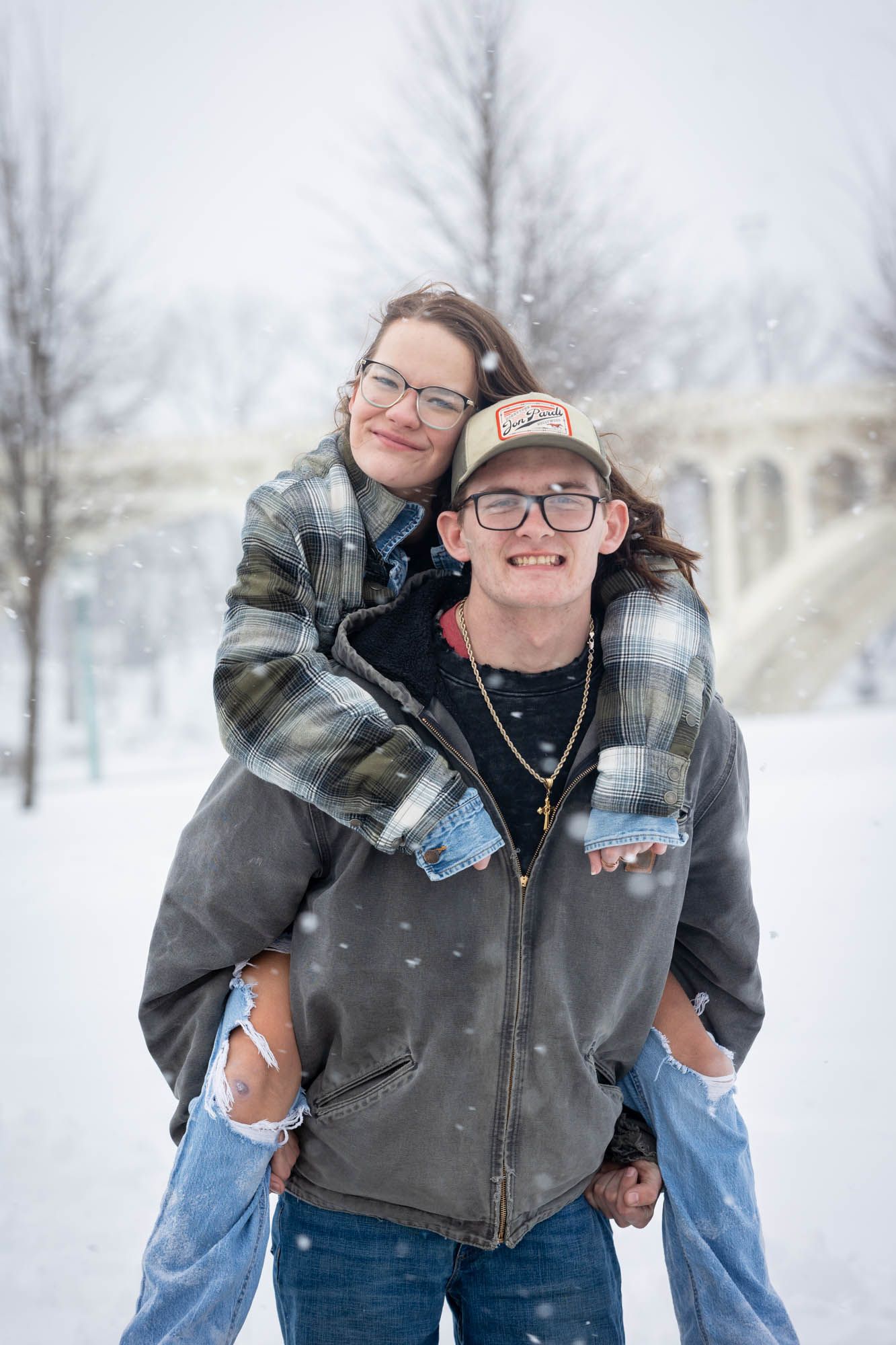 Young couple posing in snowy winter scene, woman on mans back, both wearing glasses and plaid jackets.