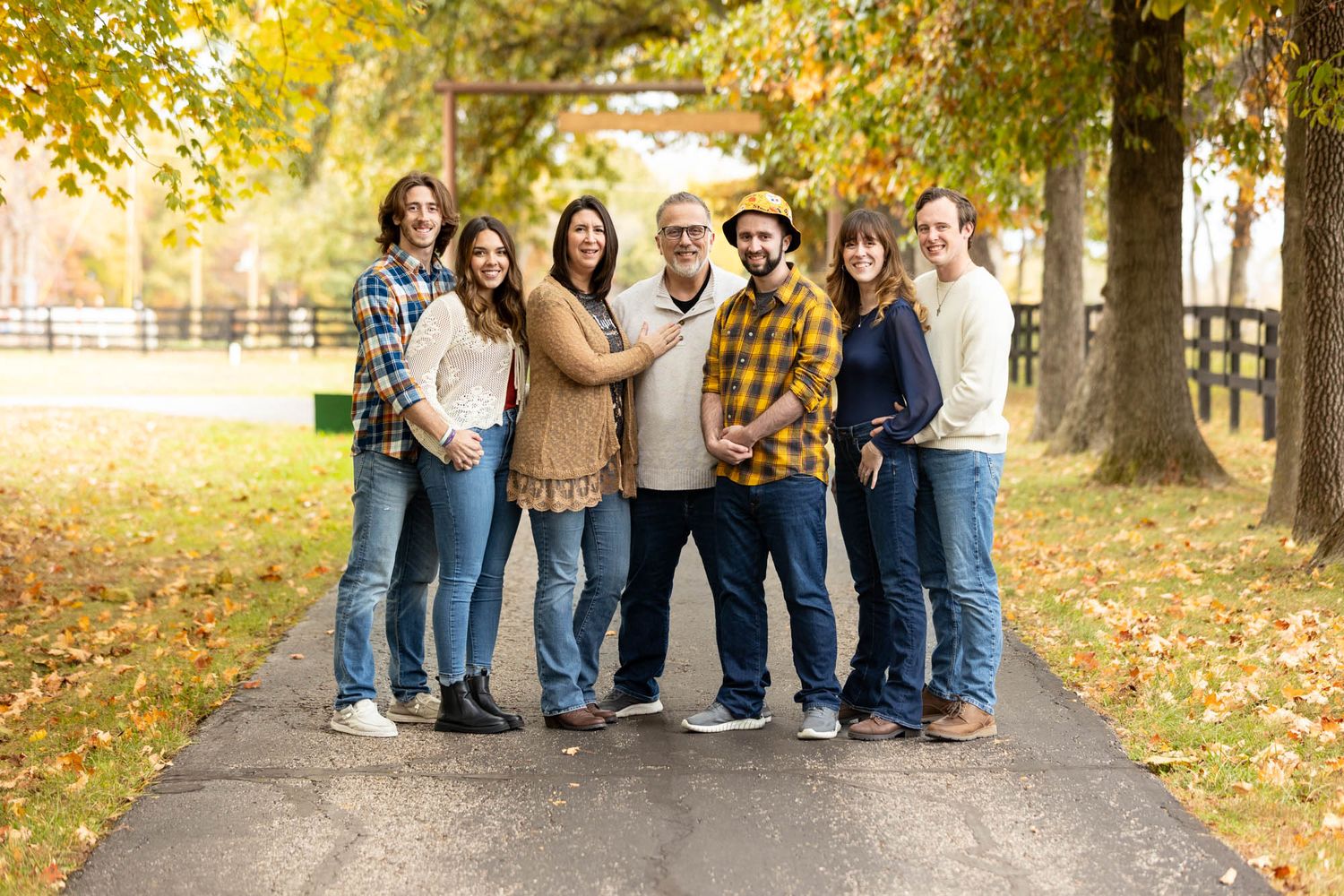 Happy family of seven smiles together on a tree-lined driveway during autumn foliage season.