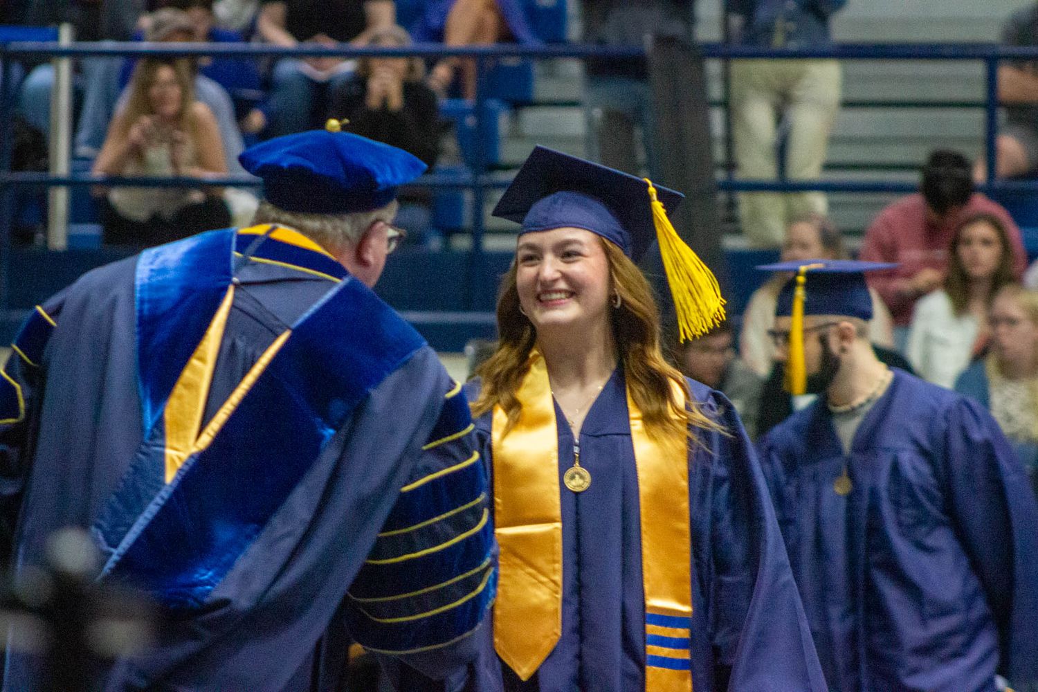 A smiling female graduate in blue cap and gown walks across the stage at a college graduation ceremony.