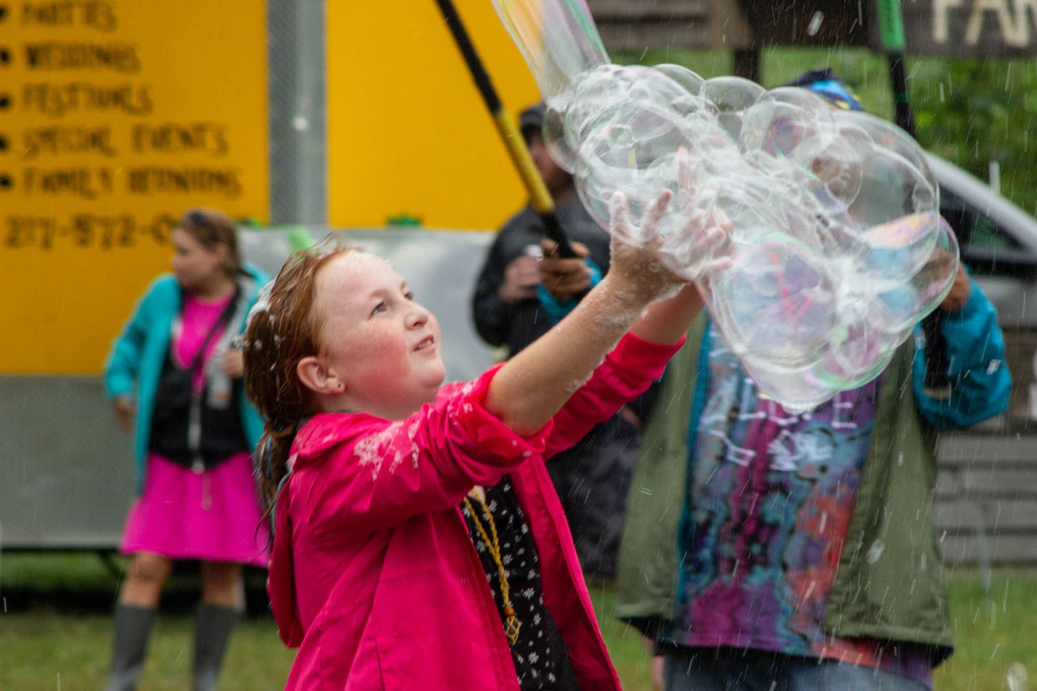 Young girl in pink jacket reaches up to catch giant soap bubbles outdoors.