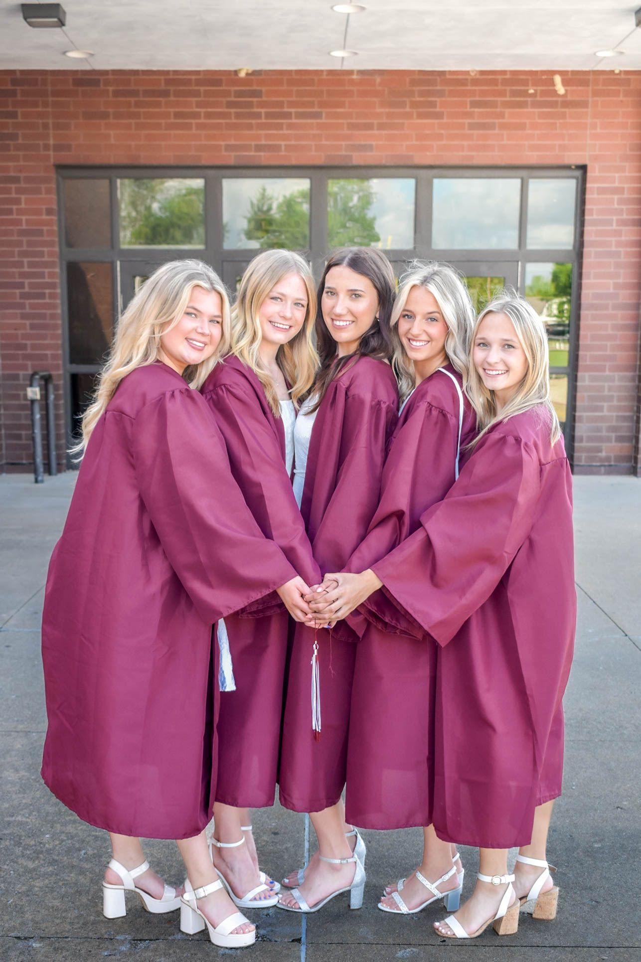 Five happy female graduates pose together wearing maroon graduation gowns outside a brick building.