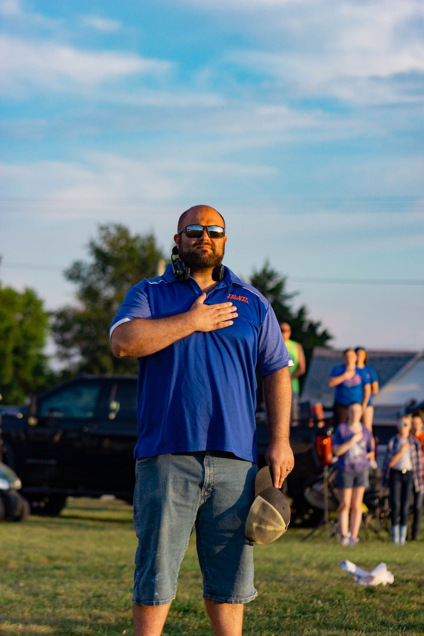 Bald man in blue polo shirt holds hat to chest during national anthem at outdoor event.