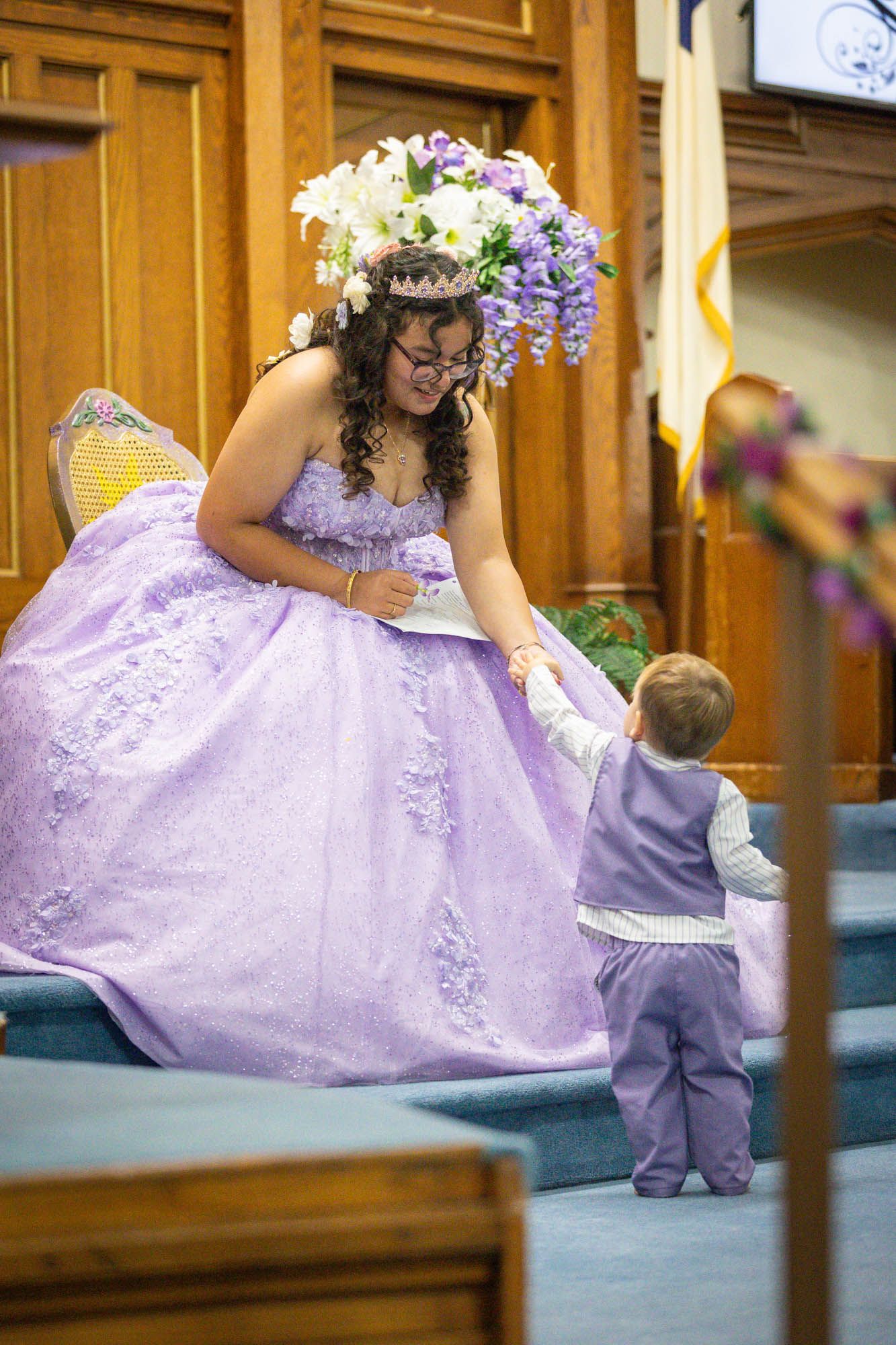 A young woman in a purple quinceañera gown with floral crown holds hands with a toddler boy in matching purple suit at church.