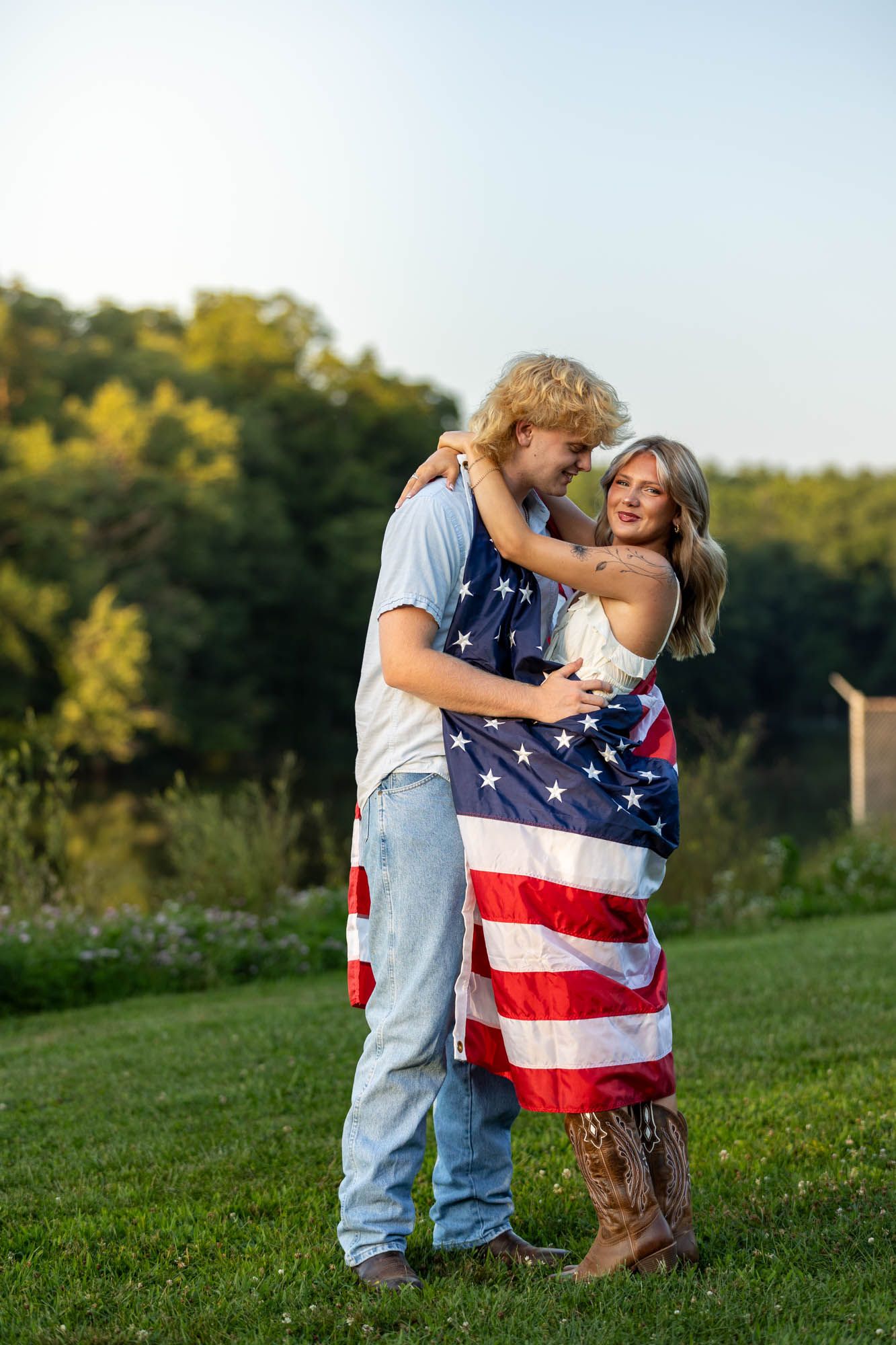 Young couple wrapped in American flag embracing outdoors on green grass with trees in background.