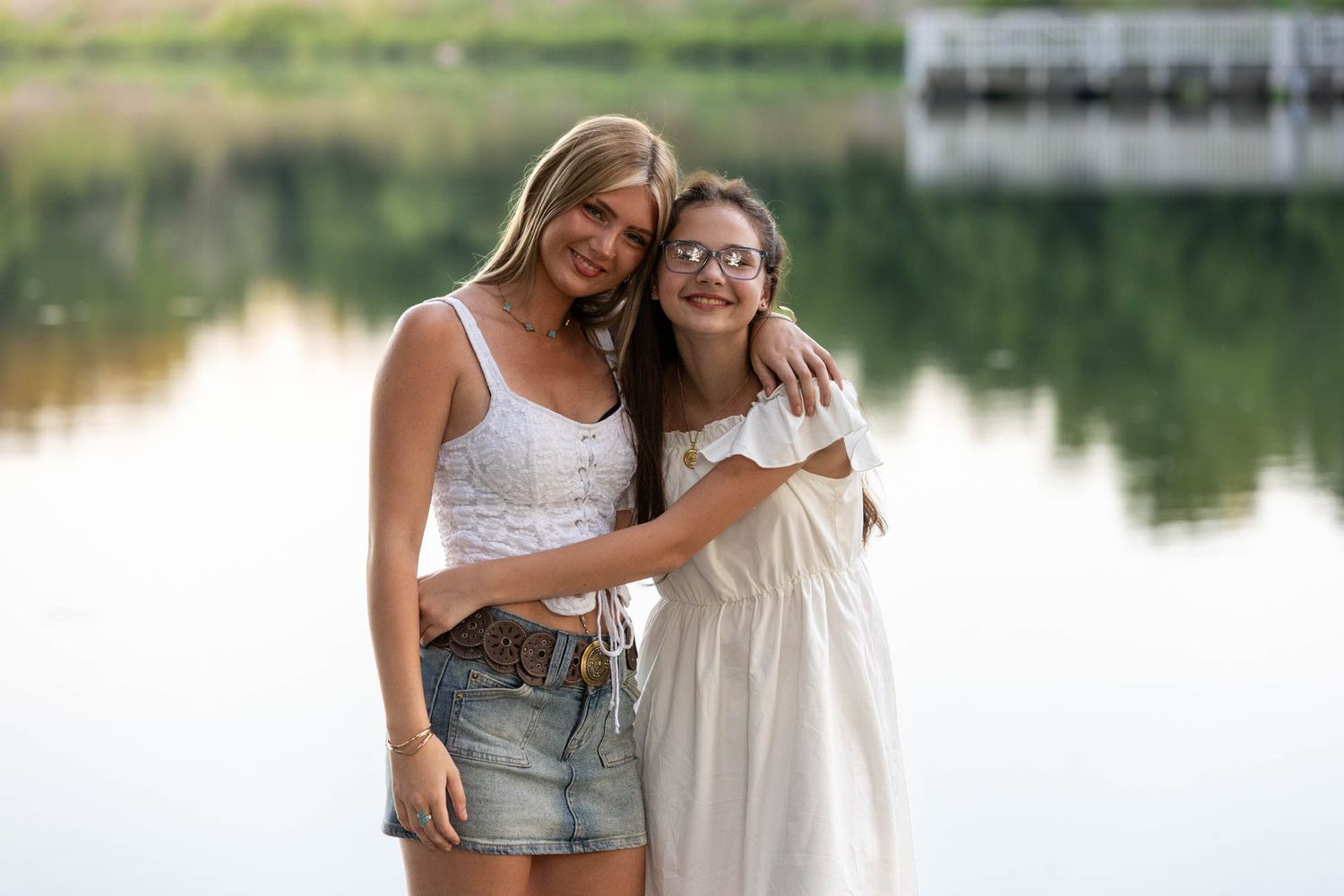 Two young women hugging and smiling together near a serene lake, one in a white dress and one in a denim skirt.