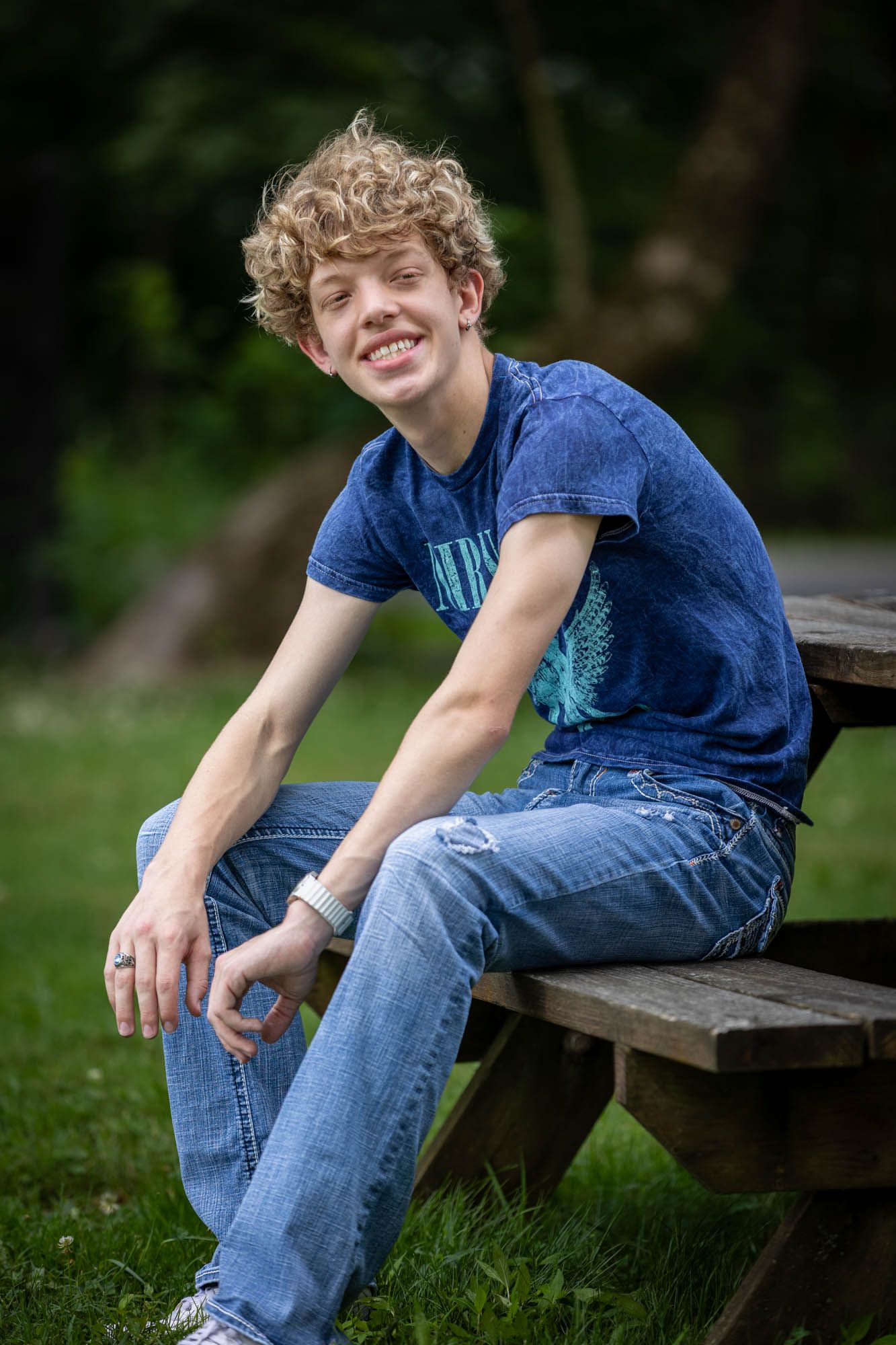 Smiling teenage boy with curly hair sitting on a wooden picnic table outdoors wearing a blue t-shirt and jeans.