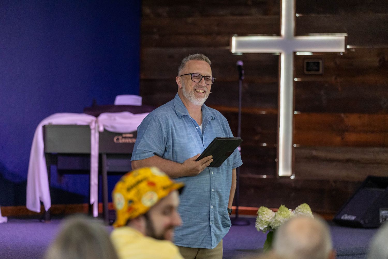 Pastor smiling and holding a book while speaking in a modern church with a illuminated cross backdrop.