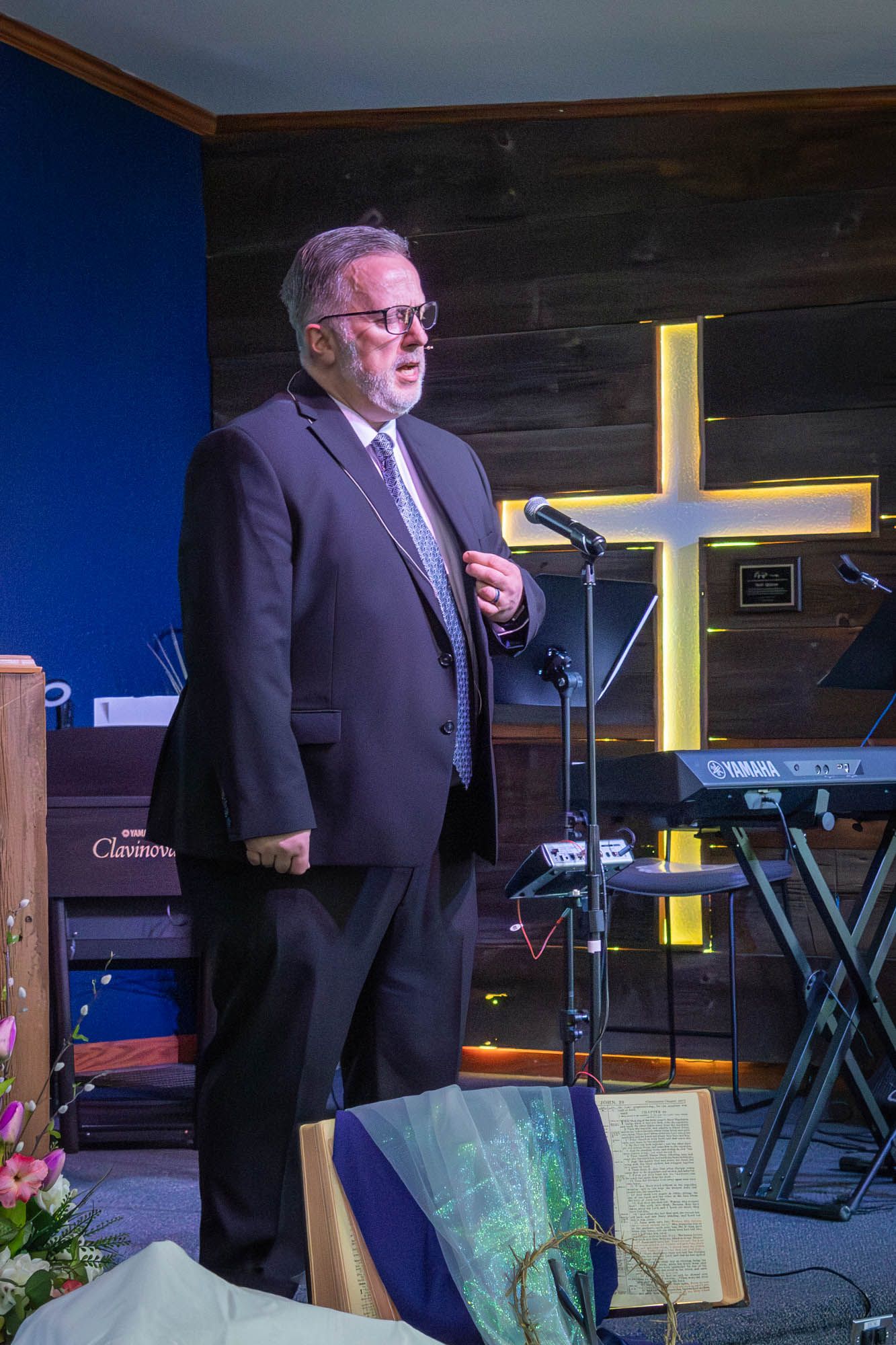 Pastor in dark suit delivering sermon at church podium with illuminated cross on wooden wall.