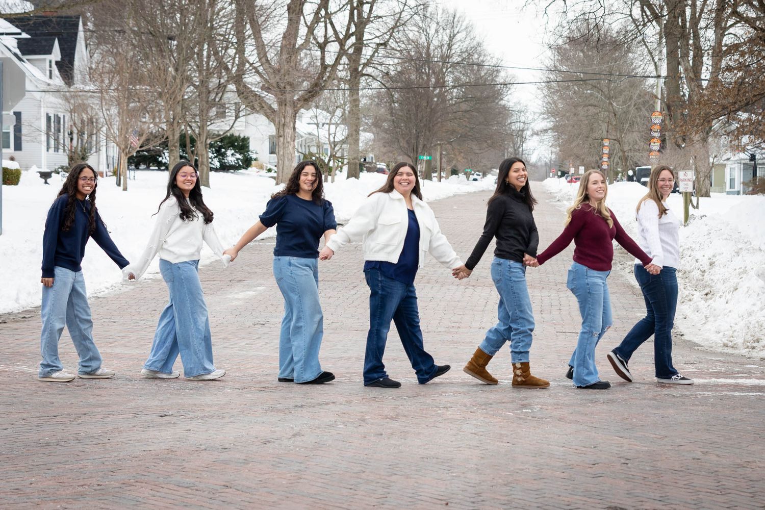 Seven young women hold hands walking in a line through a snowy winter street scene.