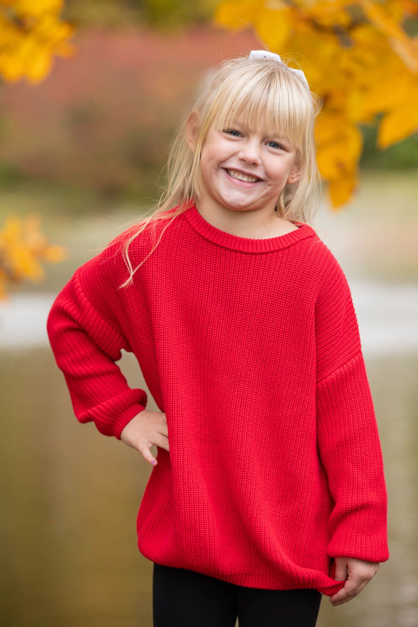 Smiling young blonde girl in an oversized red sweater posing outdoors with colorful autumn leaves behind her.