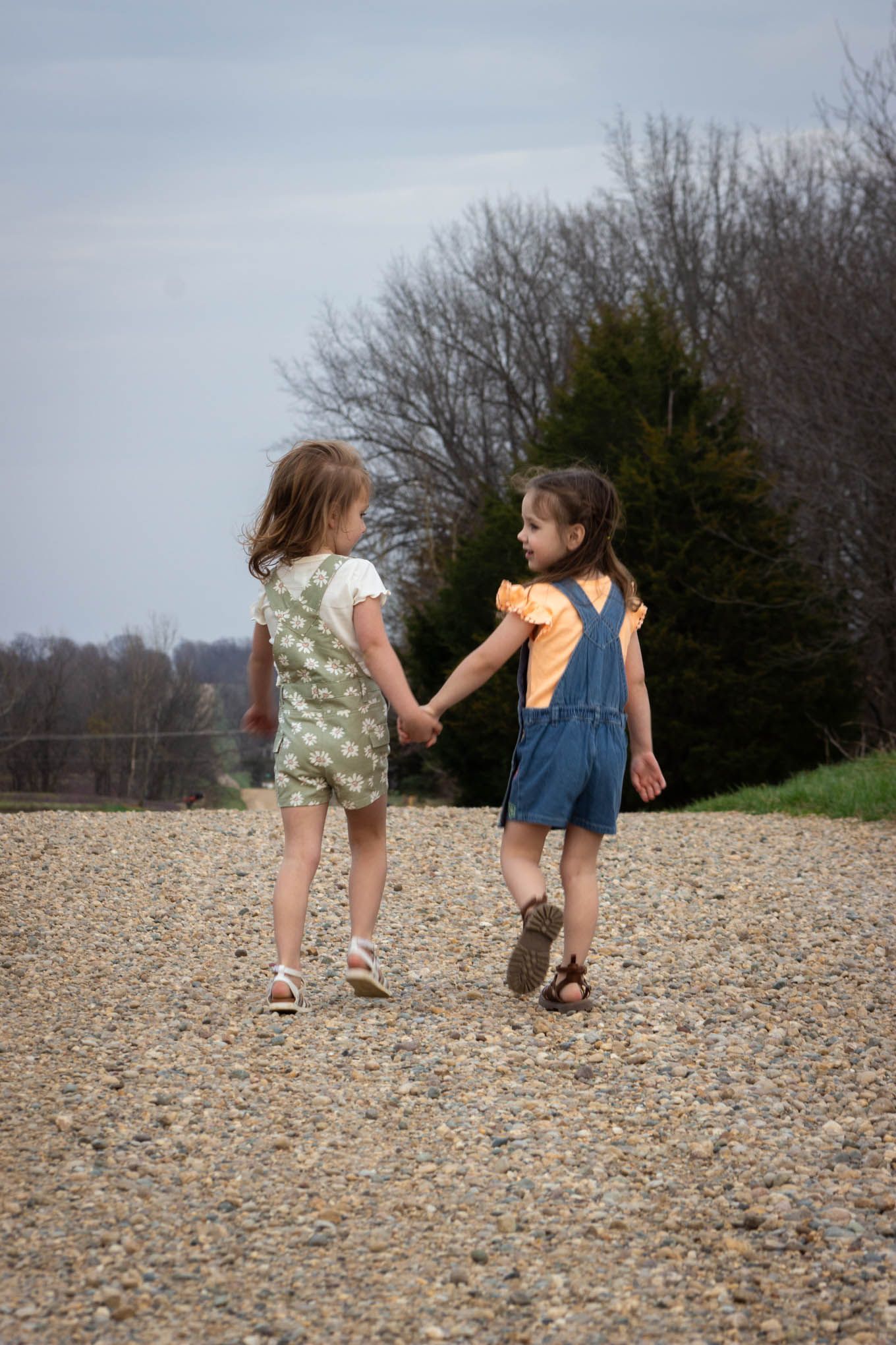 Two young girls holding hands walking away down a gravel path surrounded by trees on a cloudy day.