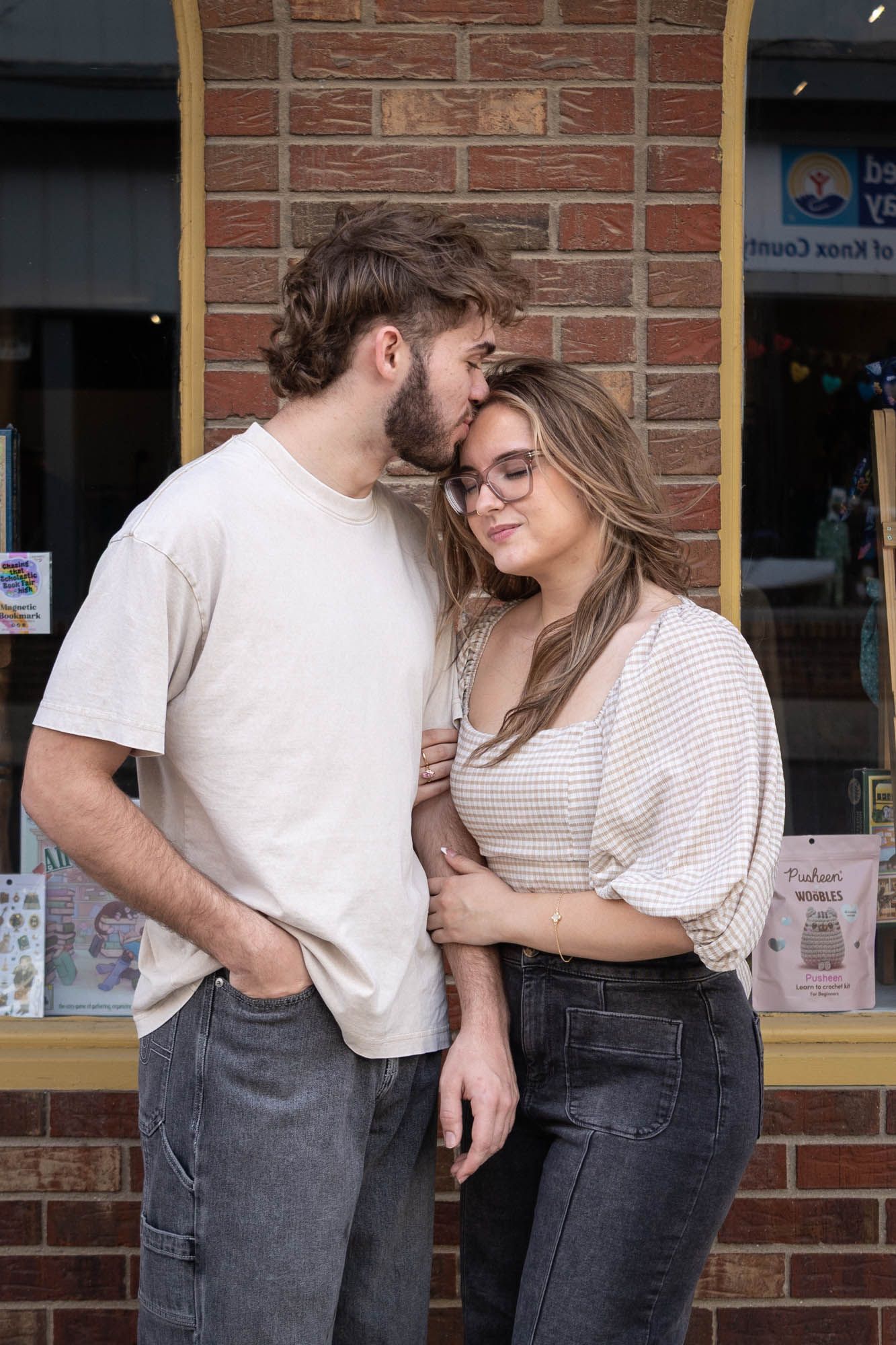 A young couple shares a tender moment as the man kisses the womans head outside a brick storefront.