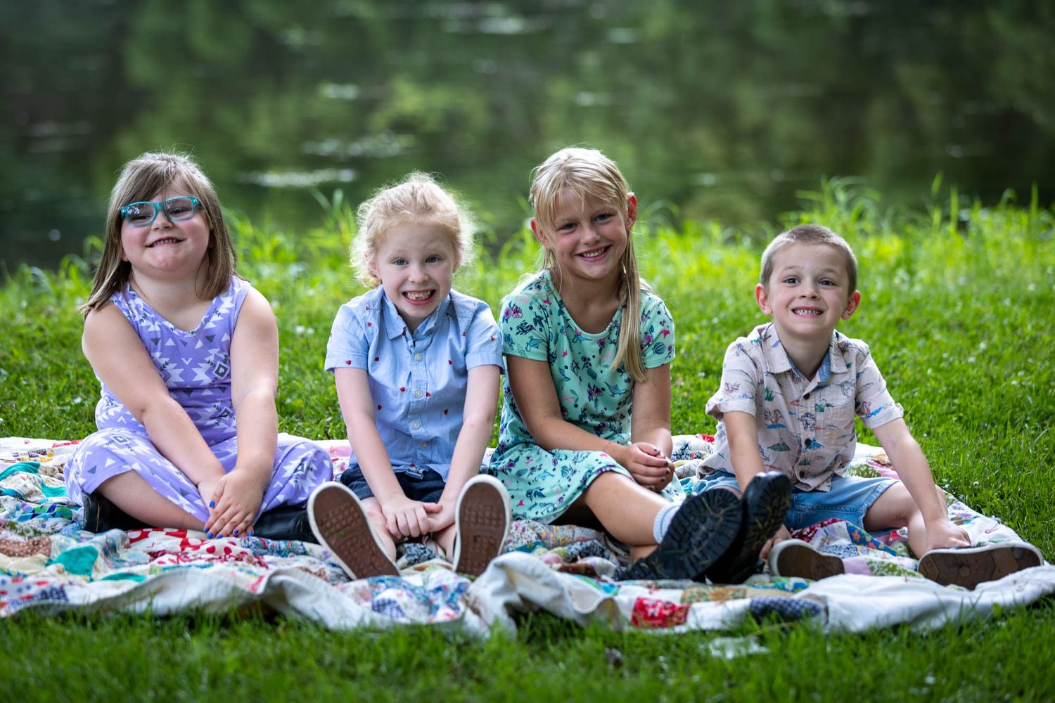 Four happy children sitting on a blanket in a park near a pond, smiling outdoors on a sunny day.