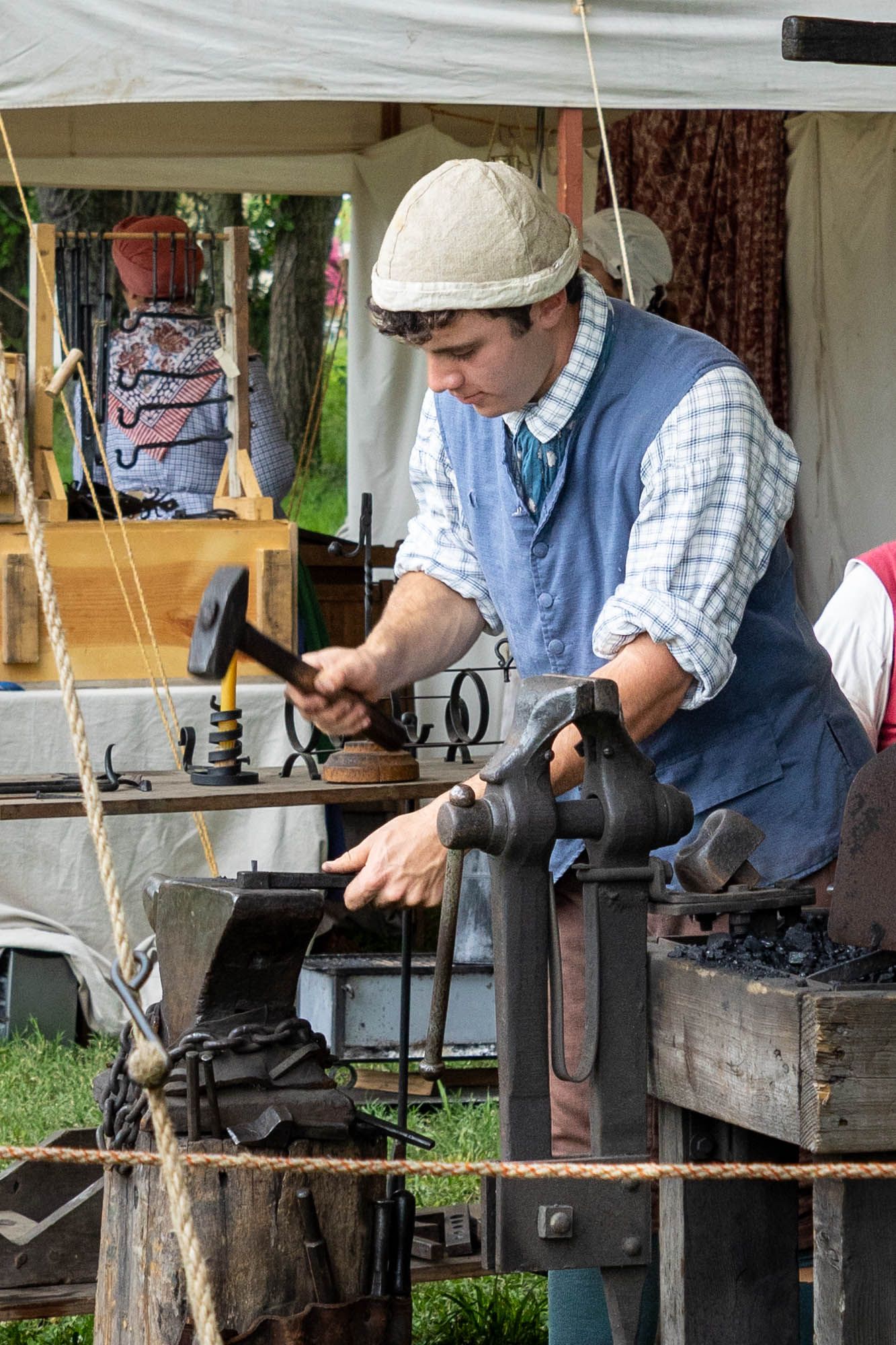 A craftsman in period clothing uses a hammer at a blacksmith demonstration at an outdoor historical reenactment event.