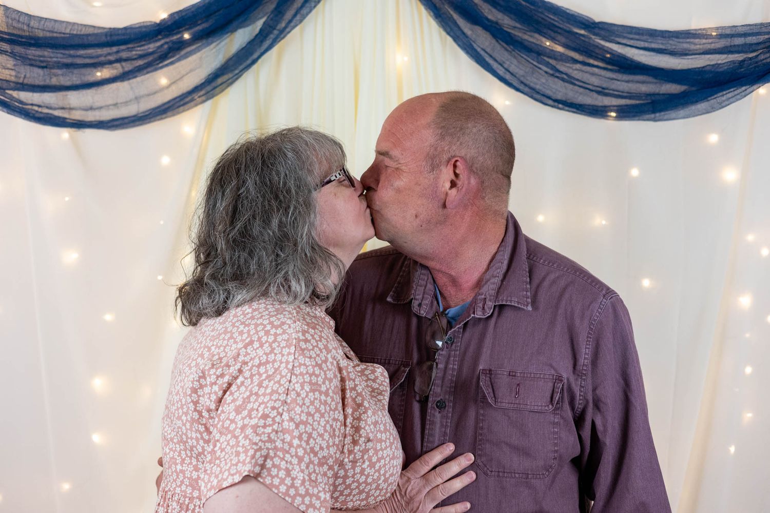 A middle-aged couple shares a romantic kiss in front of a decorative backdrop with twinkling fairy lights.