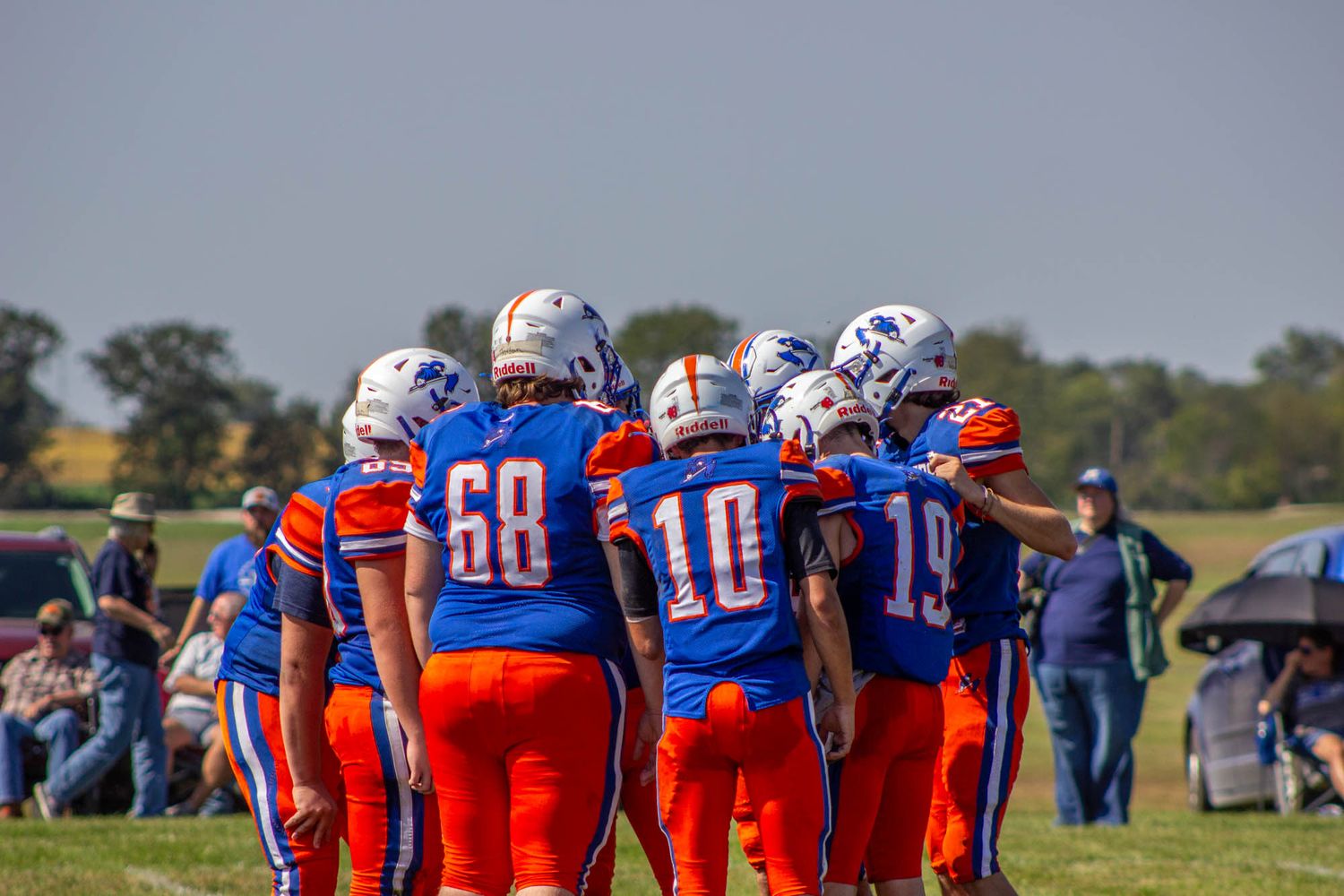 High school football team huddles together on the field wearing blue and orange uniforms with numbers 68, 10, and 19.