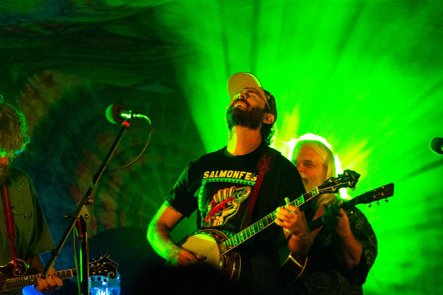 Banjo player performs on stage under vivid green lighting with bandmate beside him.