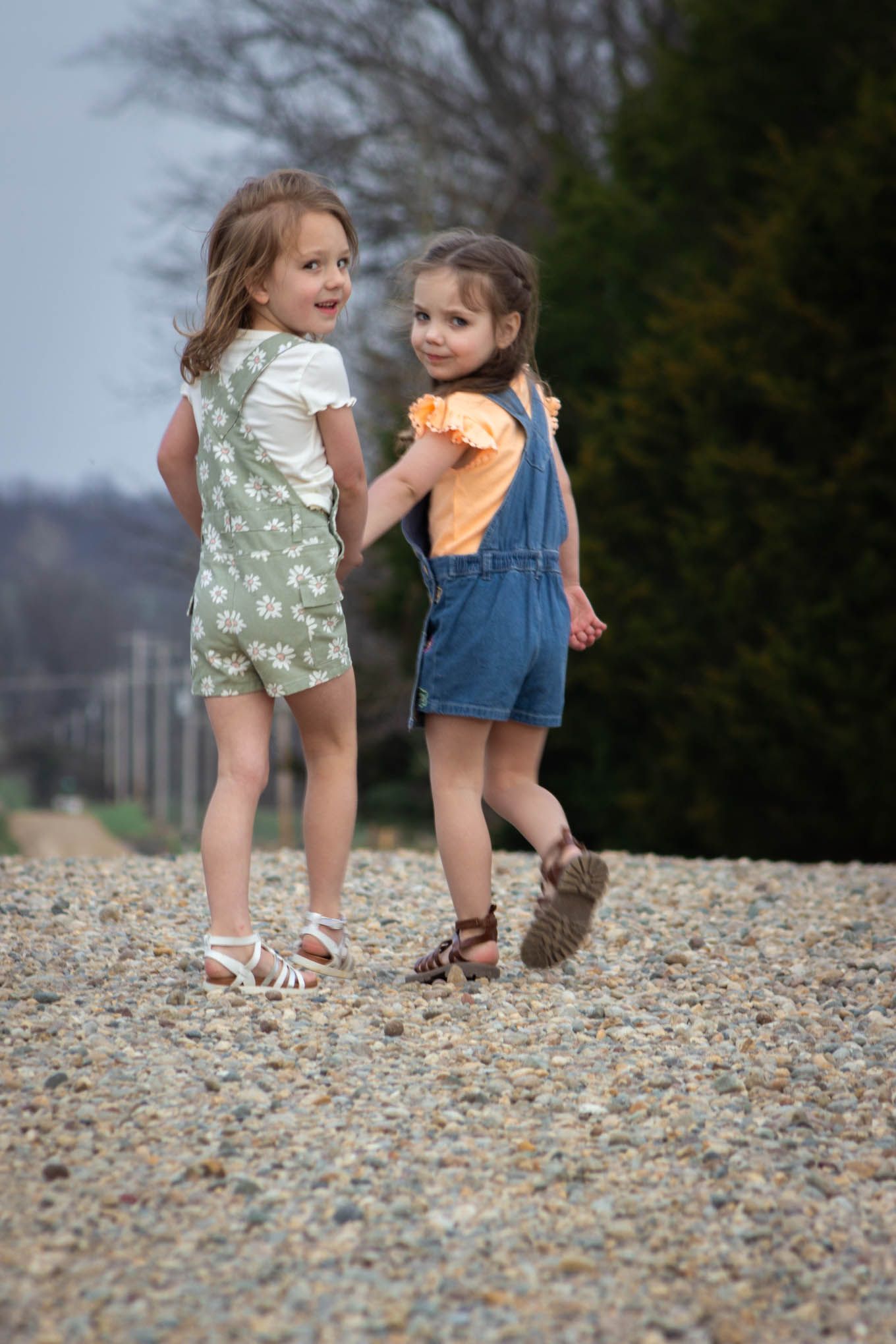 Two young girls in overalls holding hands on a gravel path, turning to look back at the camera.