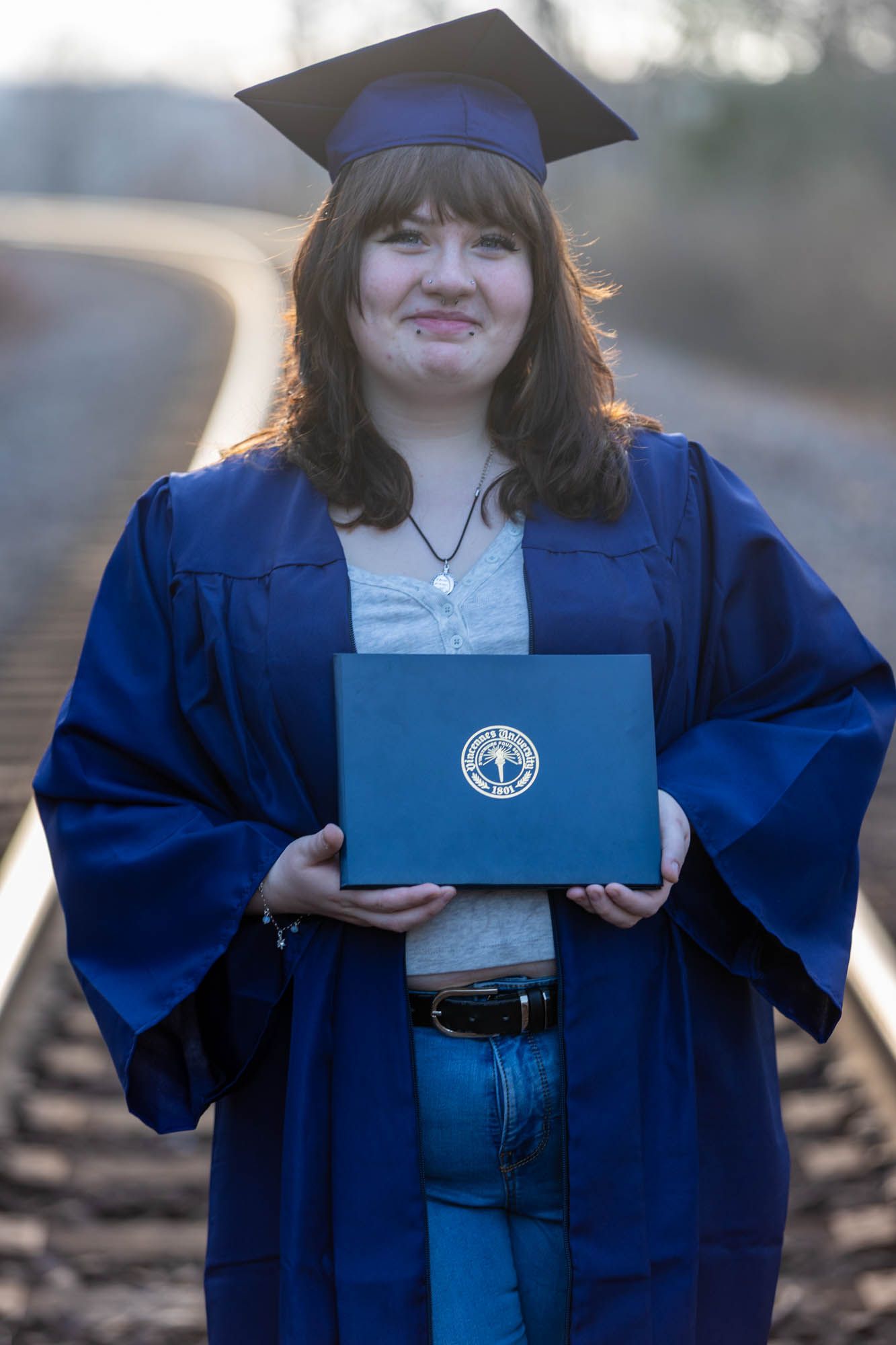 Proud graduate in blue cap and gown holds diploma while standing on railroad tracks outdoors.
