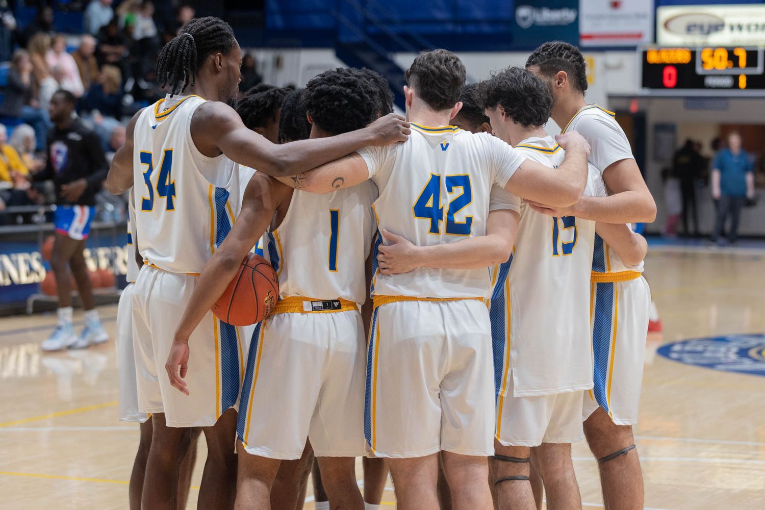 College basketball team in white and blue uniforms huddling together on indoor court before game.