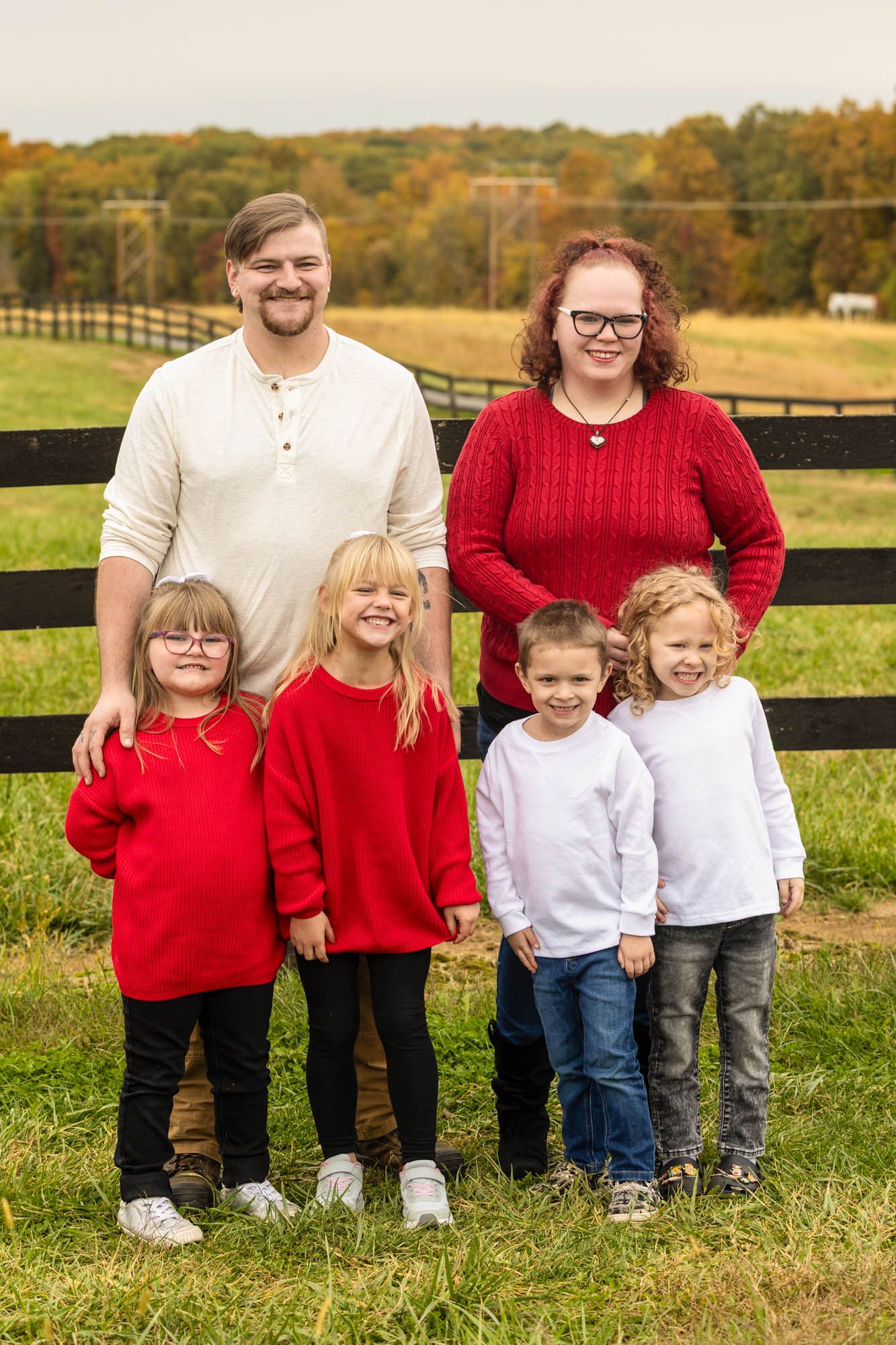Family of six with four young children posing outdoors near a wooden fence during autumn.