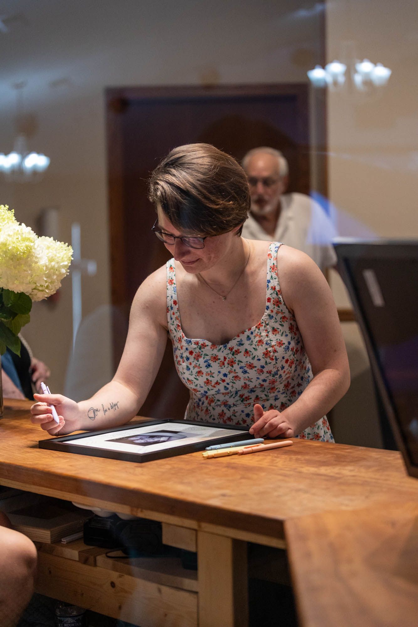 Woman in floral dress signing wedding guest book at wooden table with hydrangeas nearby.