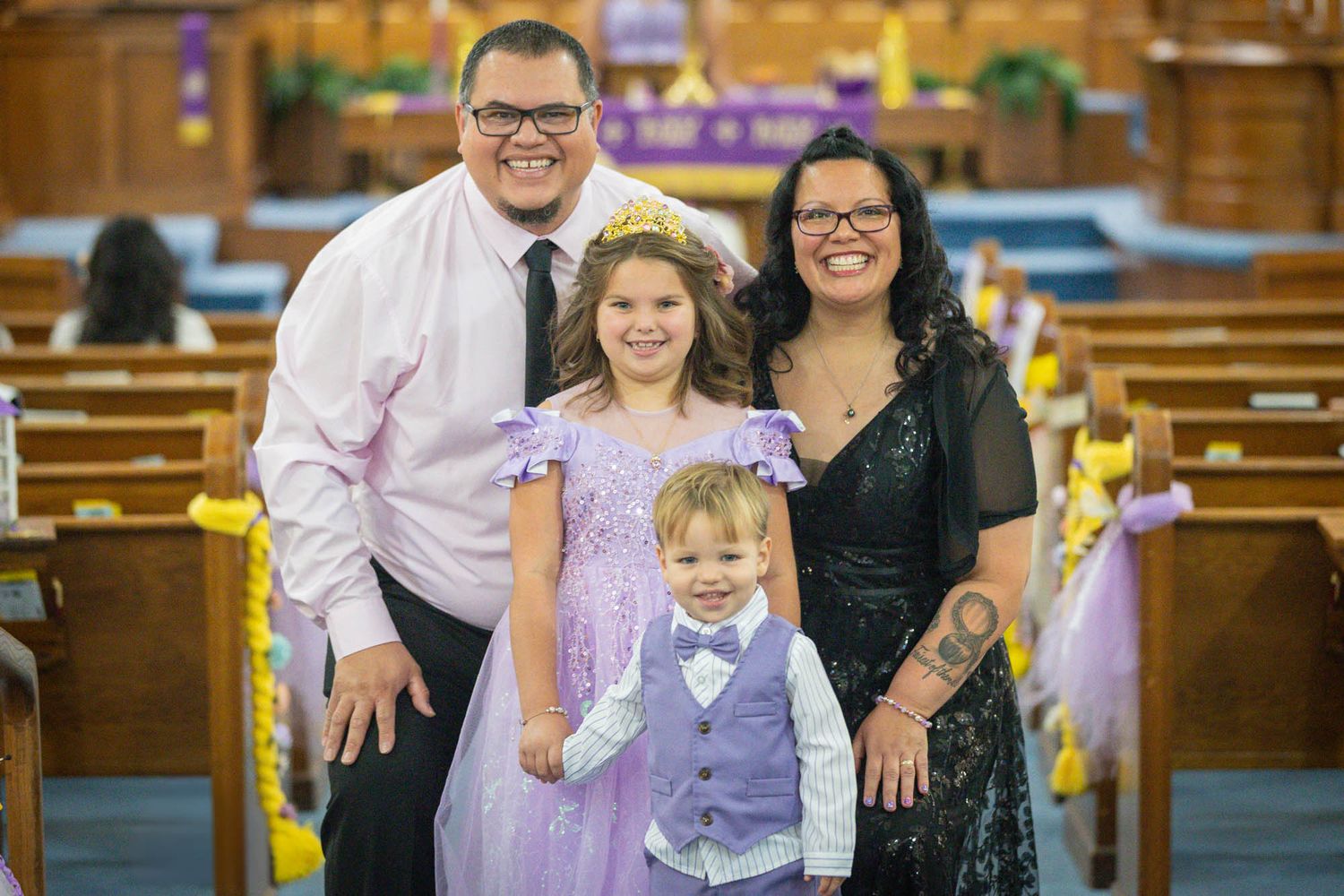 Family of four posing in a decorated church, with girl in purple dress and boy in vest, parents smiling beside them.