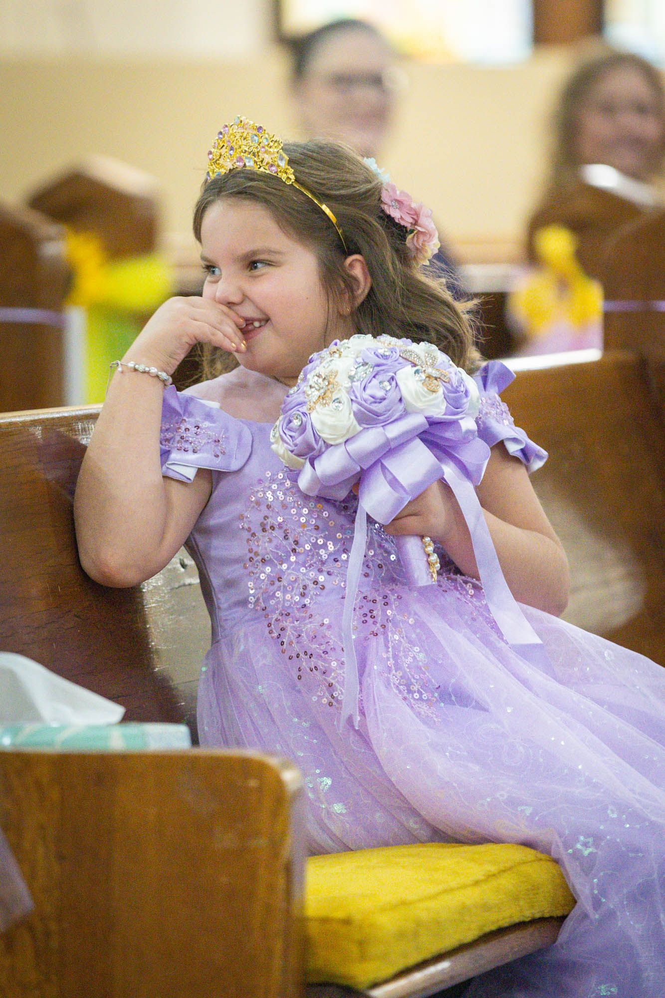 Young girl in lavender dress and gold crown sits in church pew holding purple floral bouquet at quinceañera.